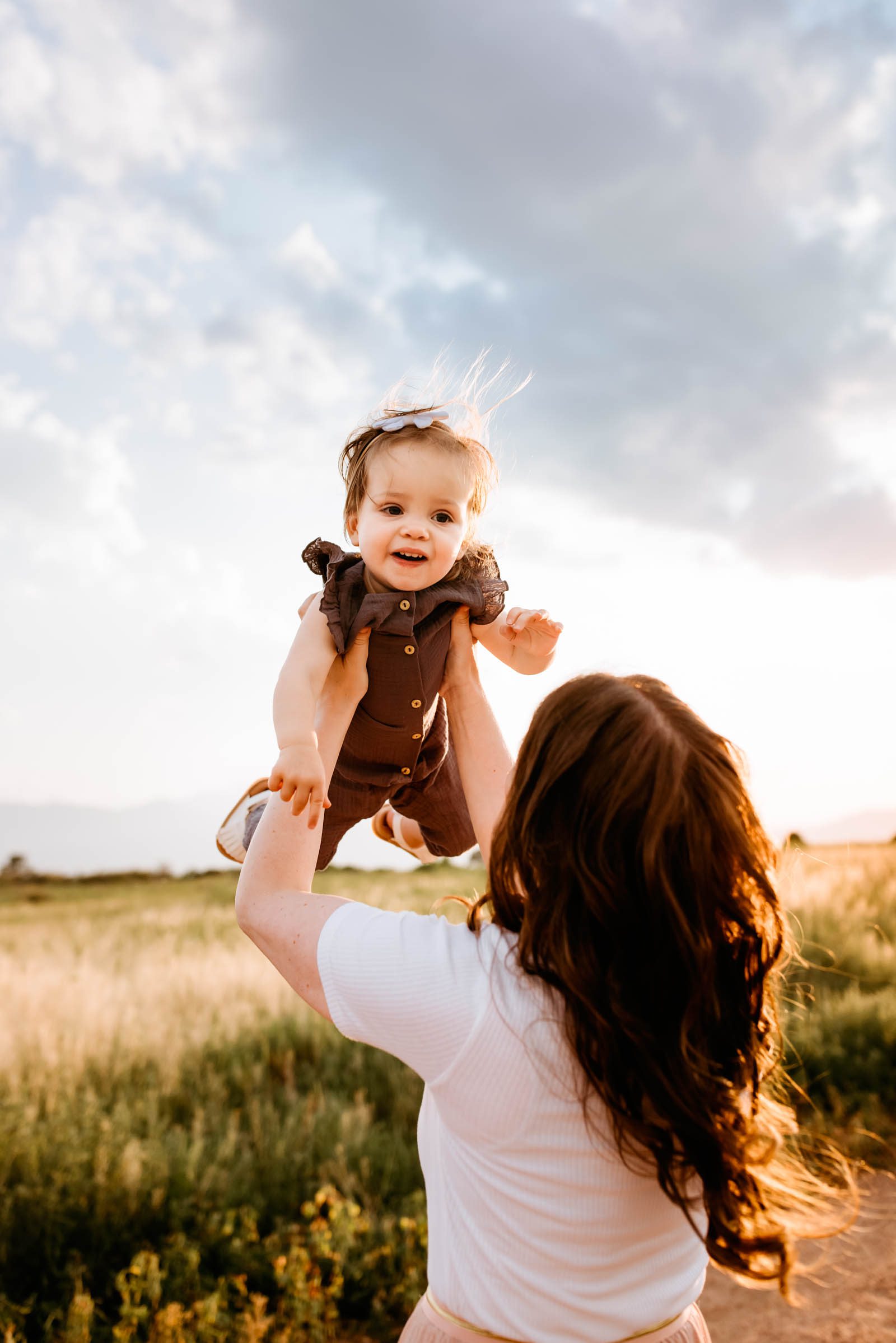 A playful moment where a smiling toddler is lifted lovingly into the air by her mother, illuminated by soft golden sunlight.