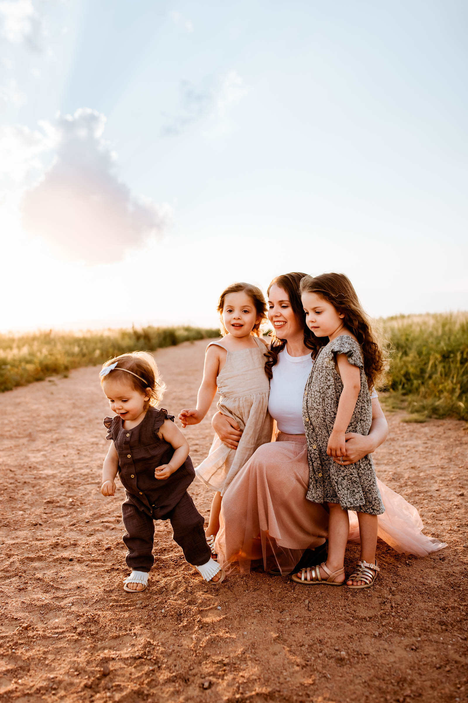 Mom lovingly surrounded by her three daughters, standing together on a scenic, sunlit path in summer.