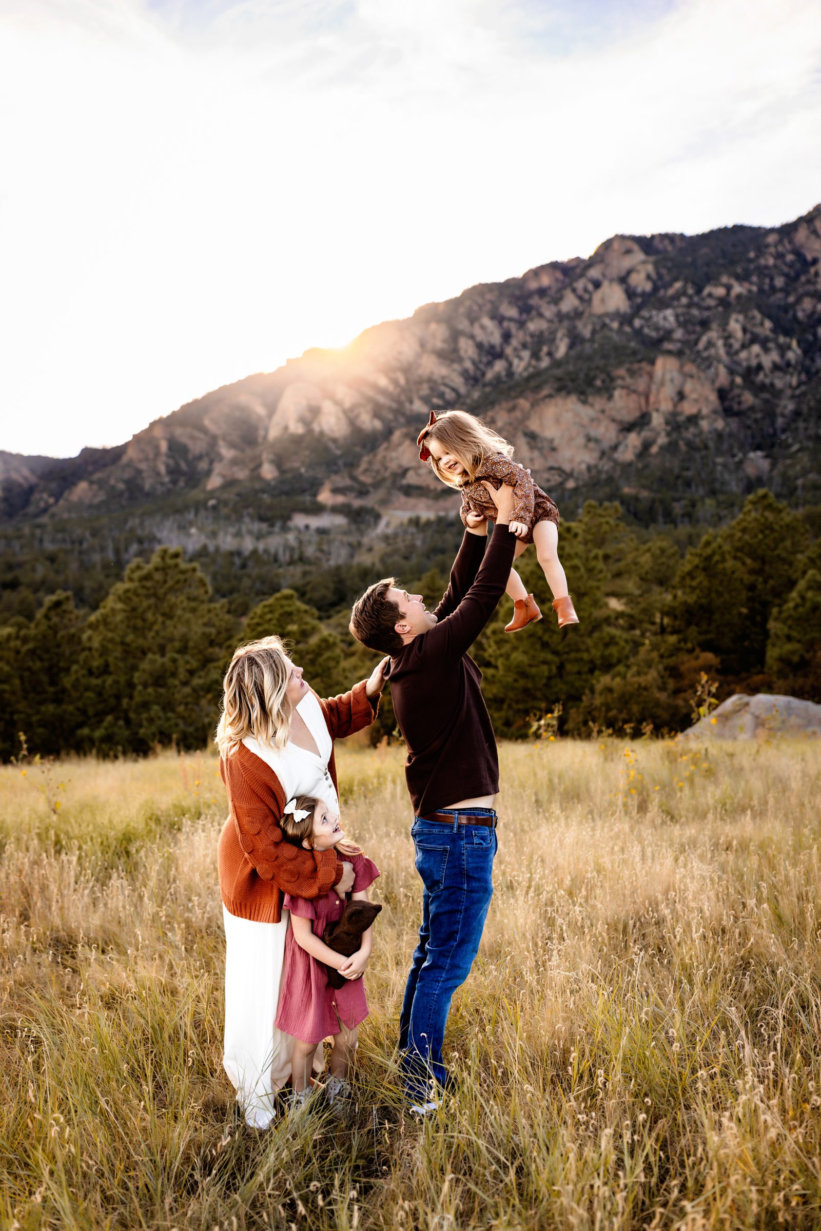 a playful family photo with dad lifting the toddler into the air while mom and big sis look up at her