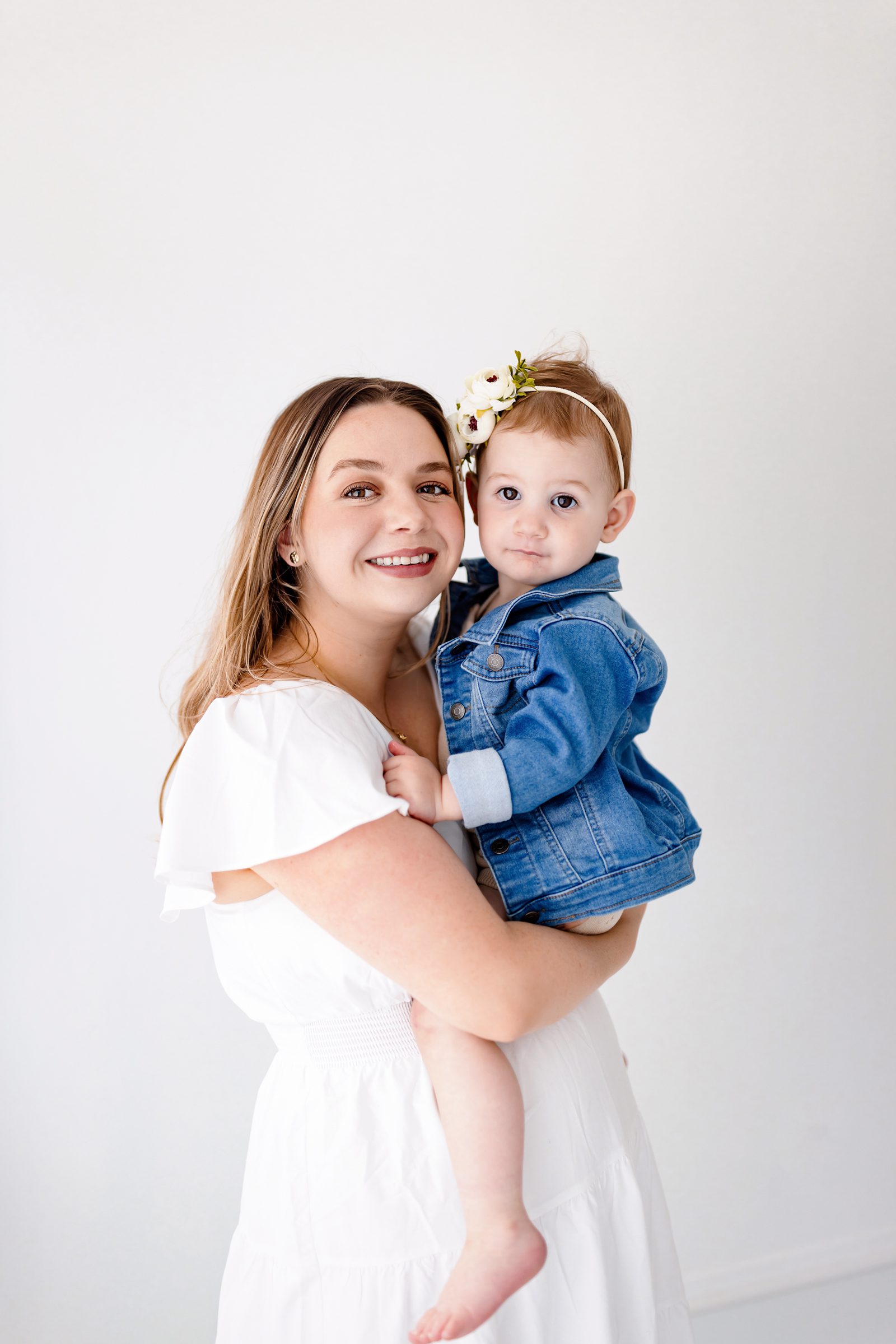 Mom holding her one-year-old in a bright white studio, both looking at the camera with soft smiles