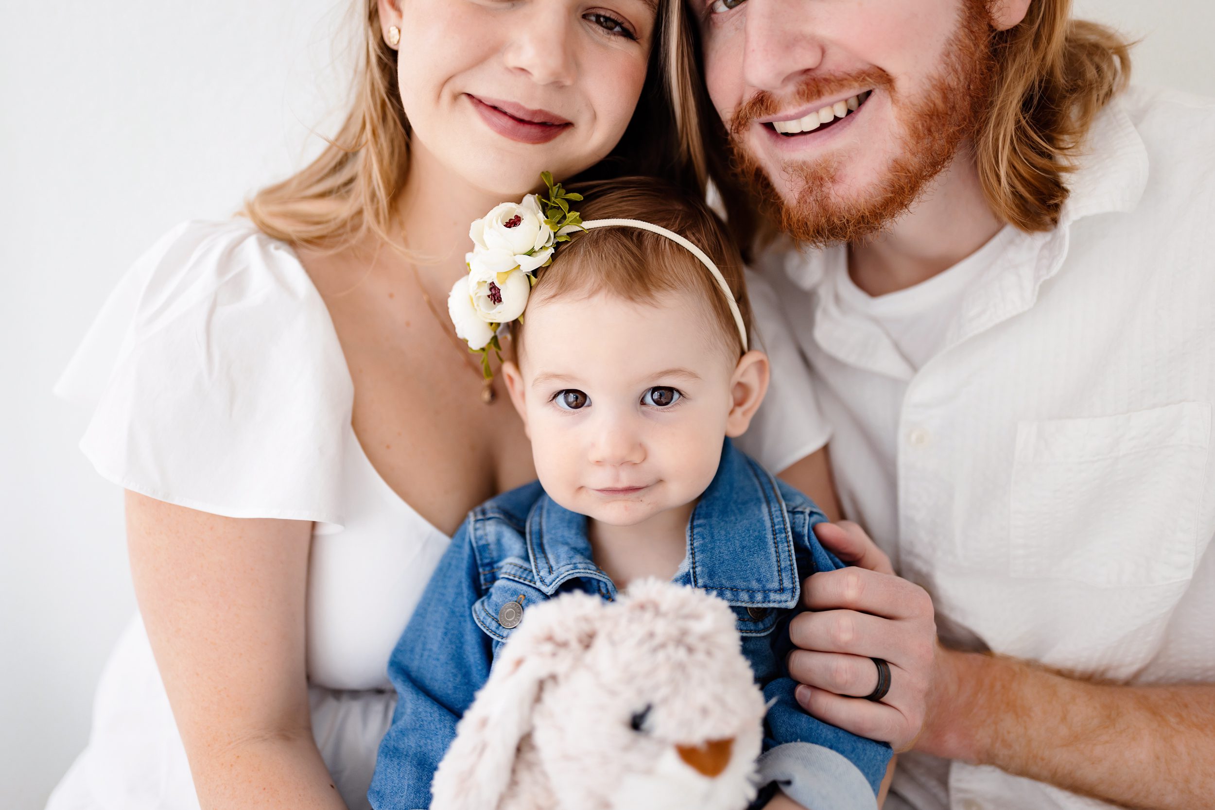 Close-up of a toddler sitting between parents, all smiling softly in a bright natural light studio