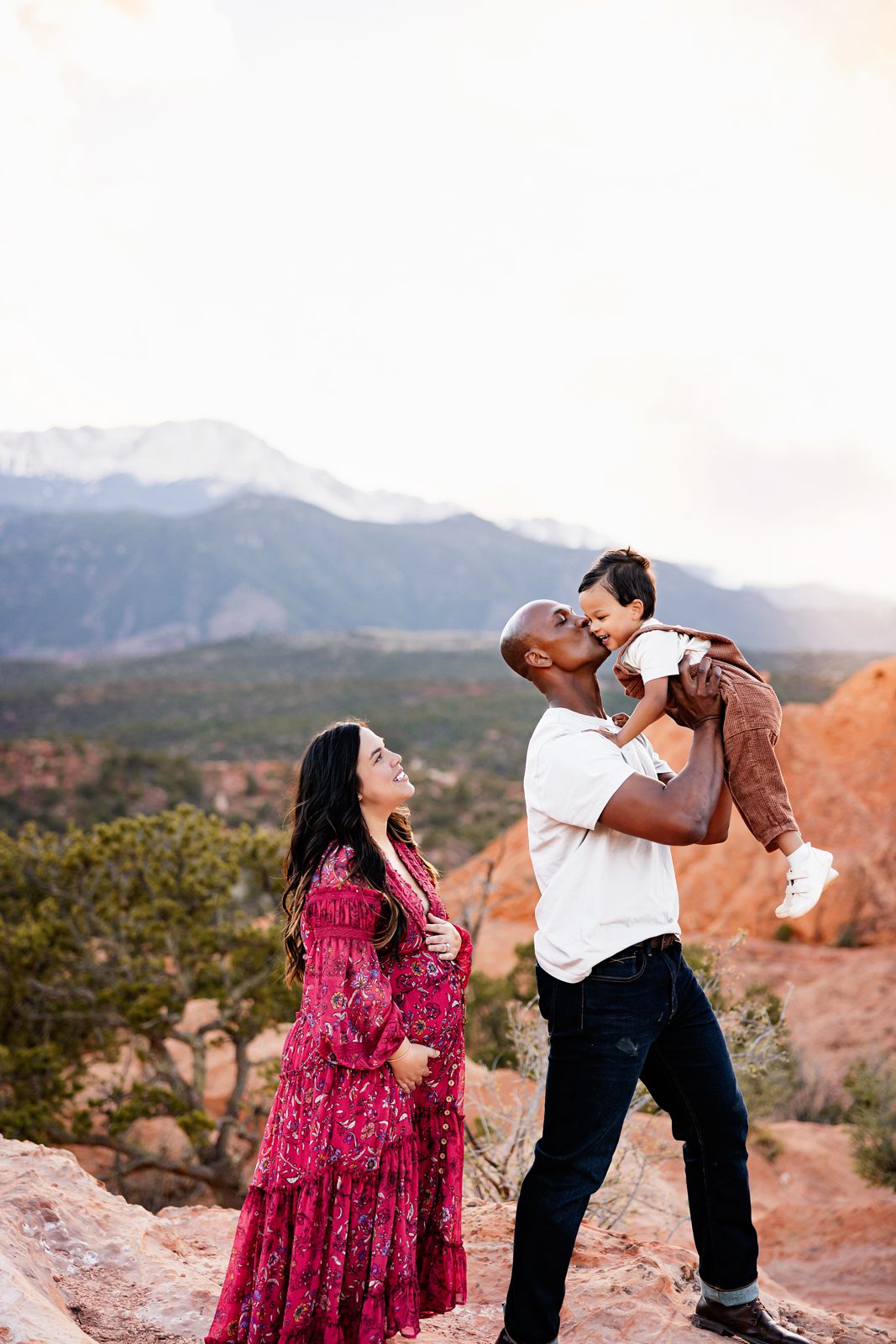 Father lifting toddler for a kiss while mom smiles nearby with scenic mountain views and soft evening light