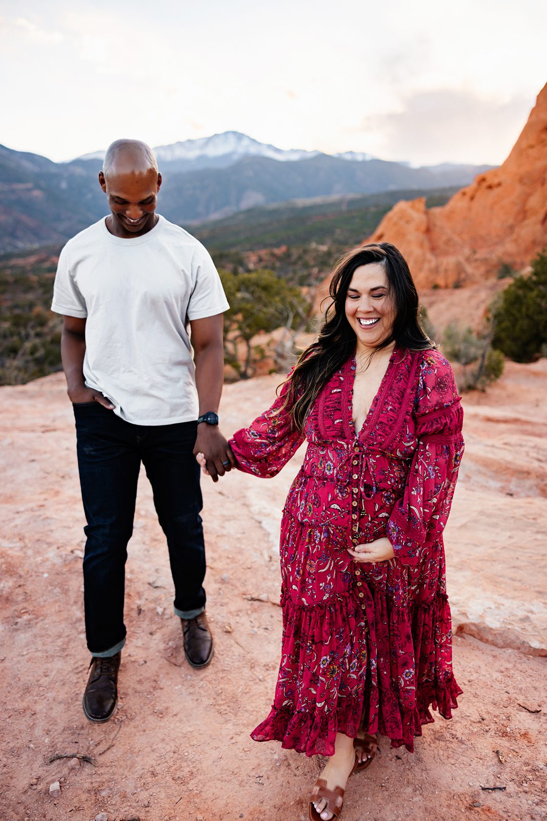 Expecting couple walking hand in hand across red rock terrain with relaxed smiles and warm sunset tones