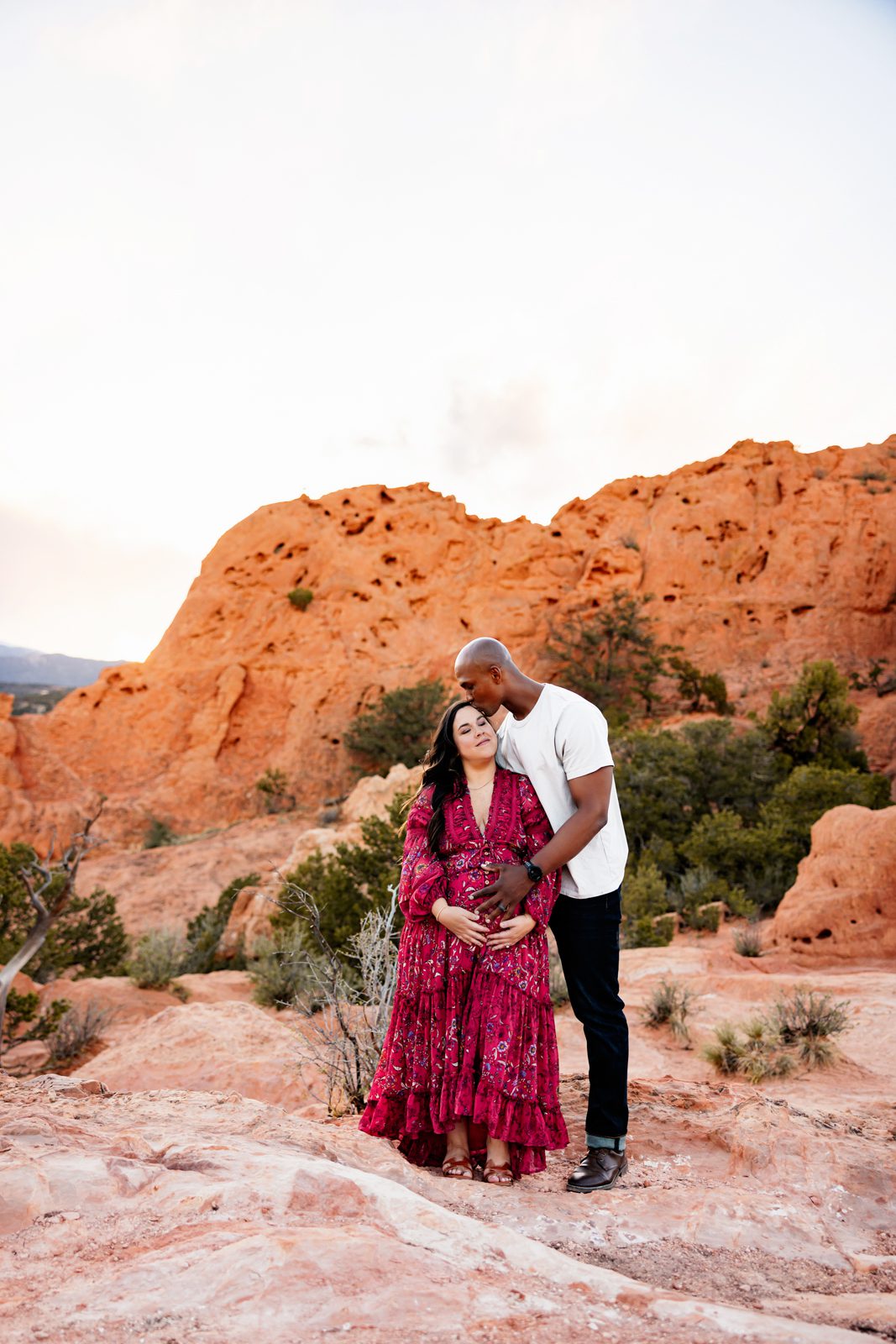 Father gently kissing mother’s head while holding her baby bump against a backdrop of red rocks and soft sky