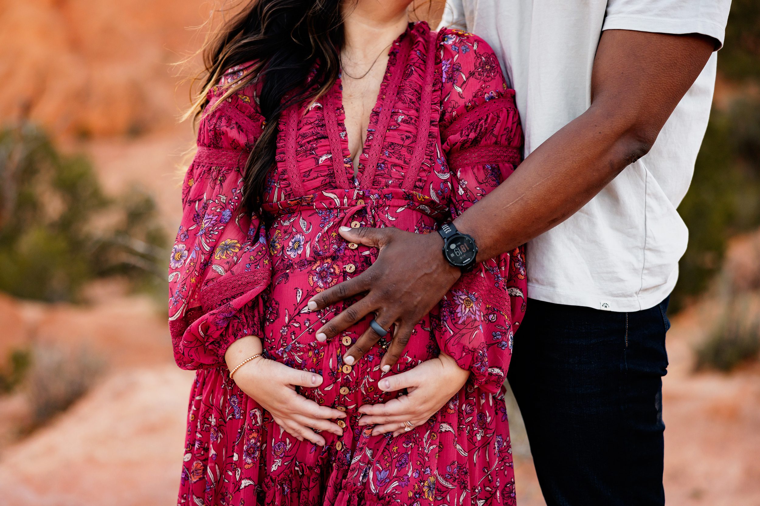 Close-up of parents’ hands resting on a baby bump with a textured red dress and soft natural light