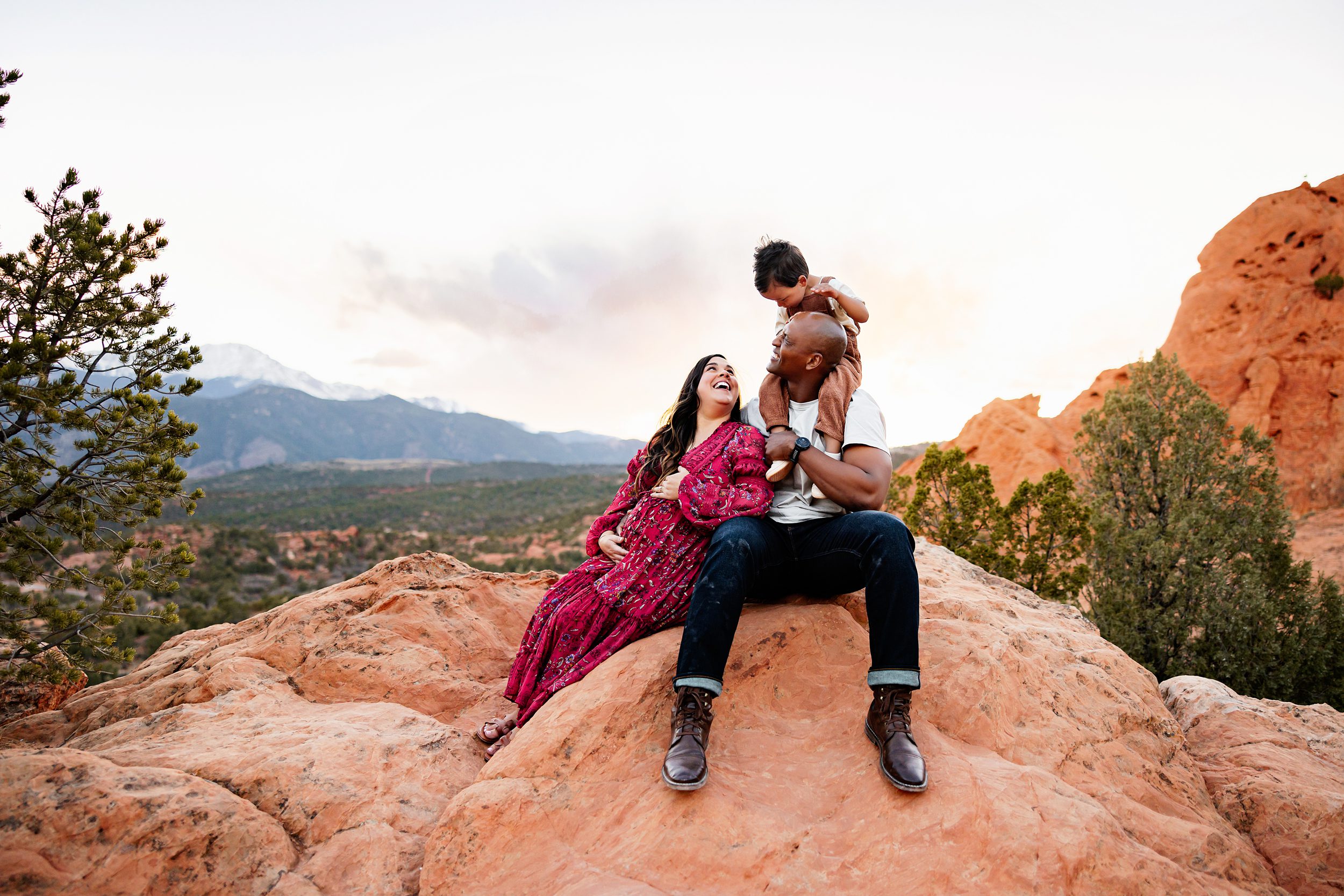 when to take maternity photos colorado featuring a family sitting on red rocks with toddler on dad’s shoulders and mountains behind