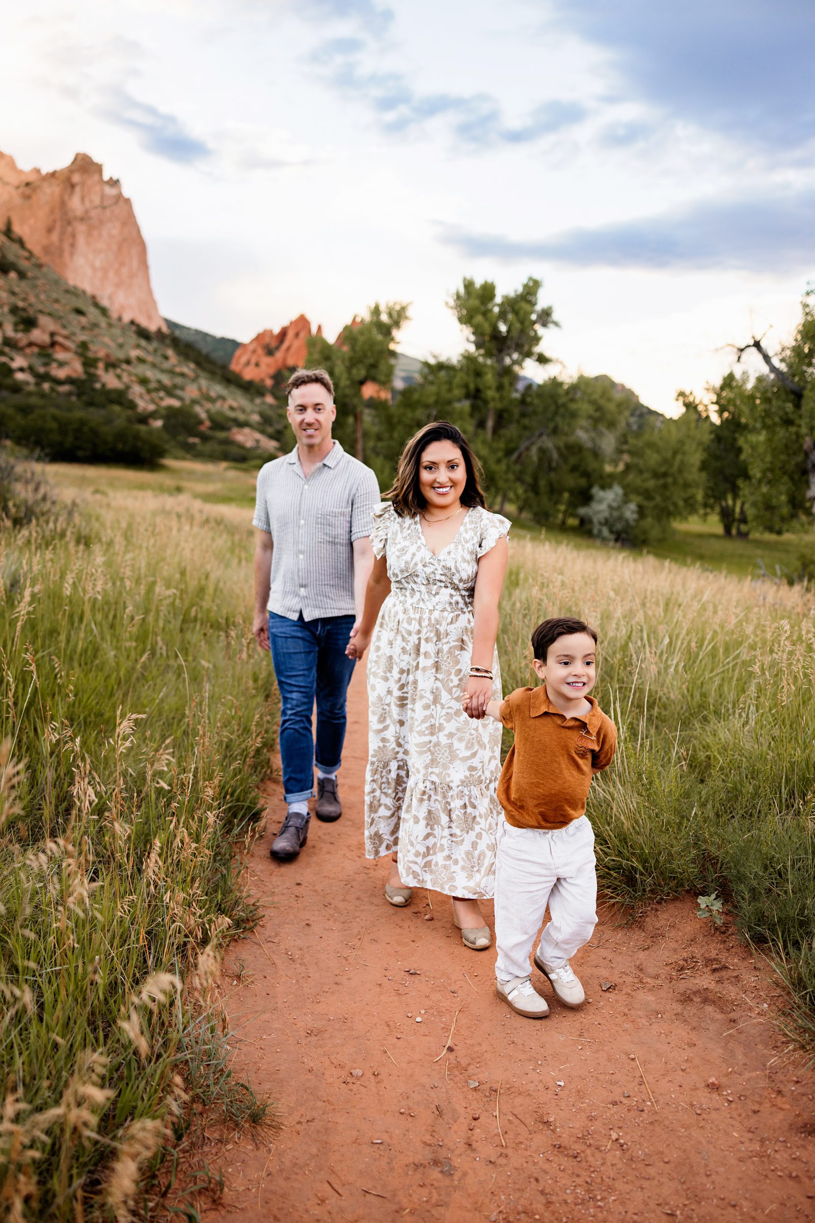 Family walking together on a dirt path with their young child leading through a grassy field at sunset