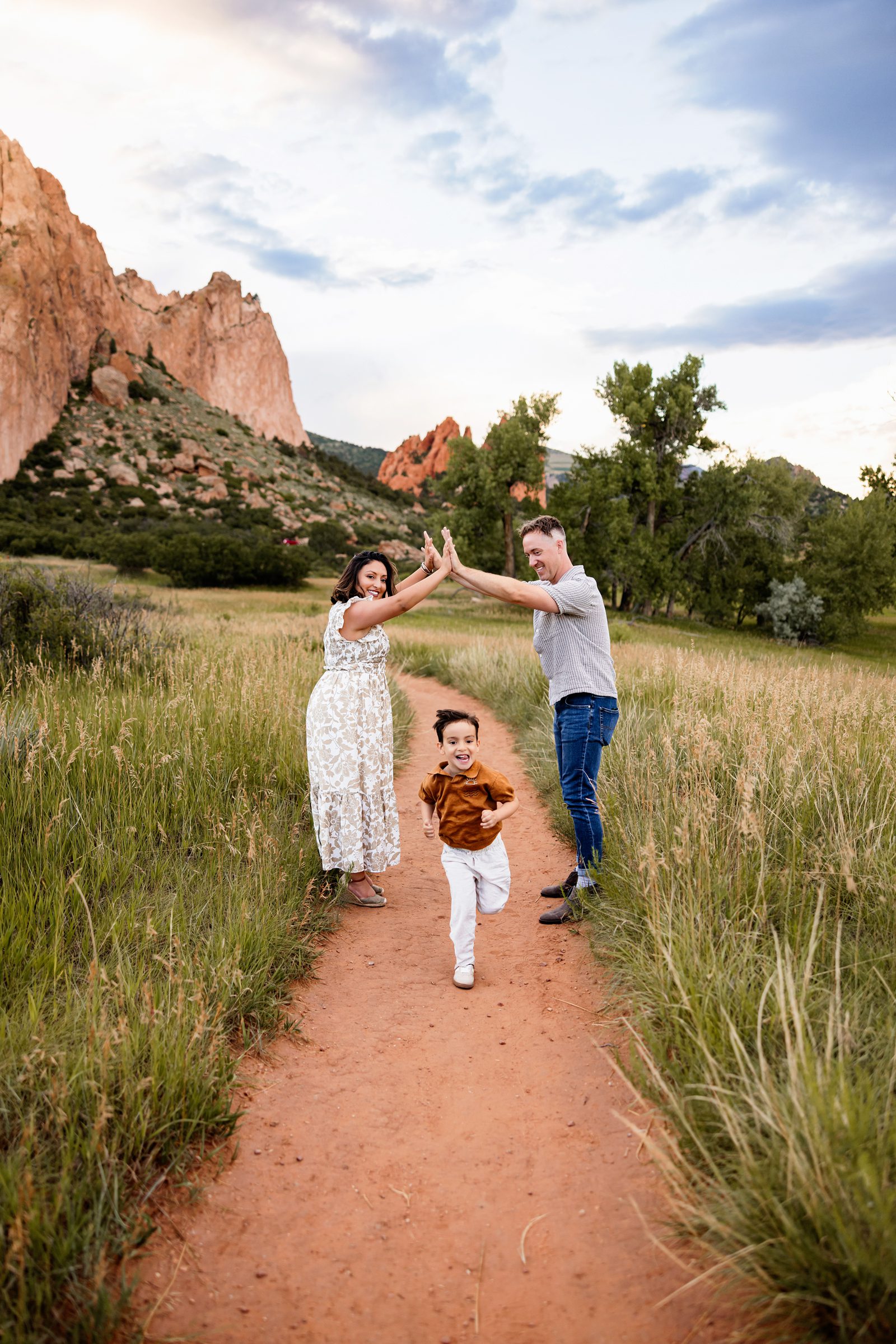 Playful family moment with a child running under parents’ raised hands on a trail with red rock formations