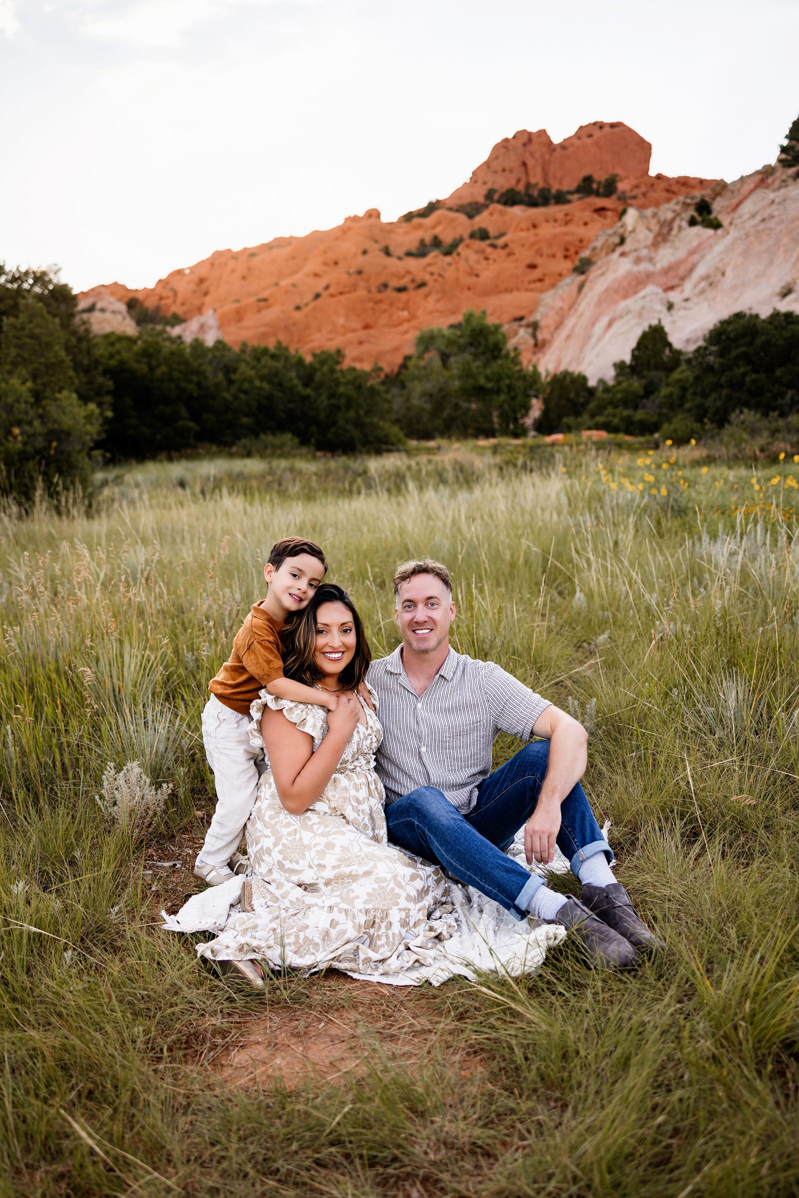 Summer Family Photos in Colorado Springs at Garden of the Gods with parents and child sitting in a meadow with red rocks behind them