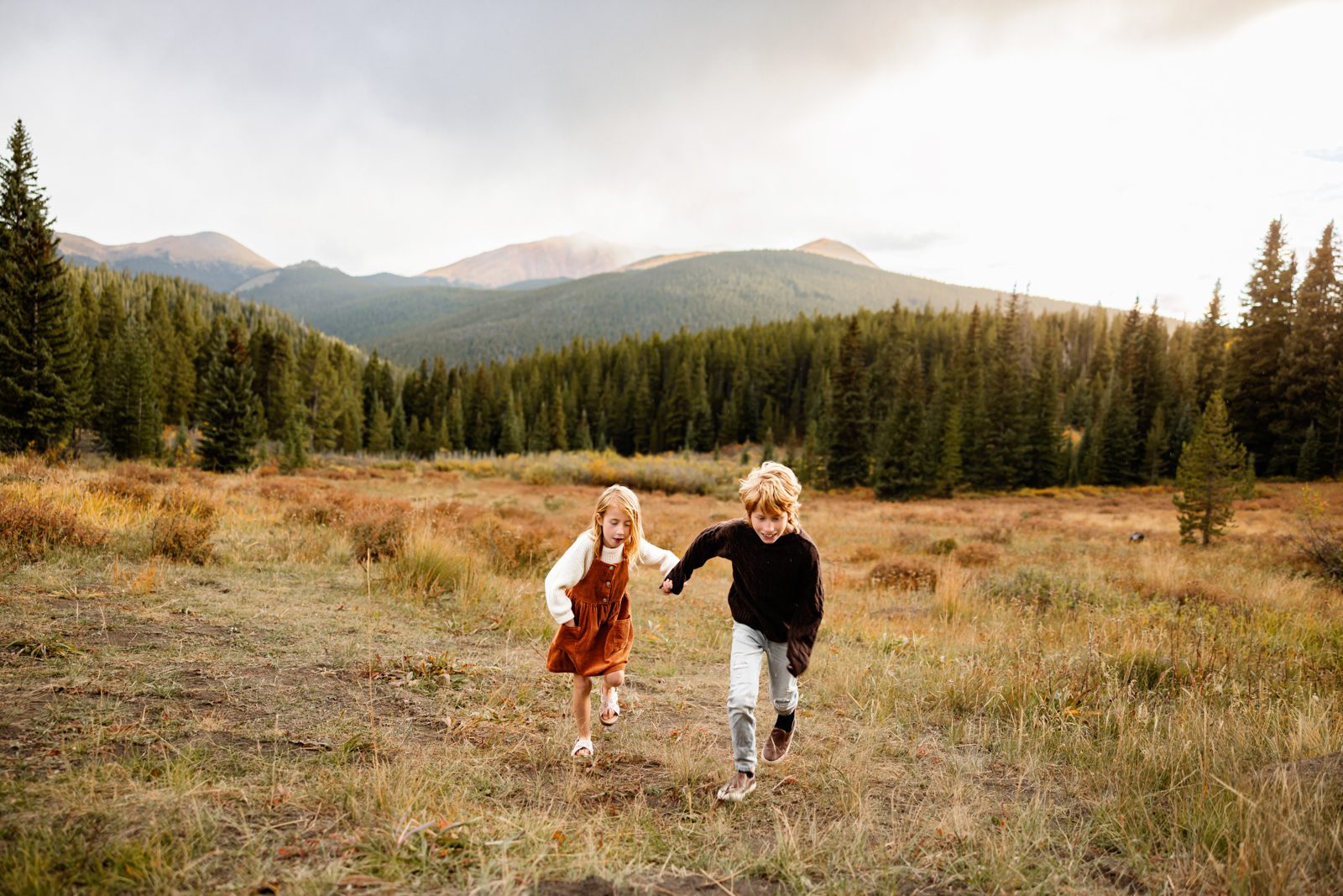 Breckenridge Family Photographer capturing siblings running through a golden mountain meadow with pine trees and peaks in the background