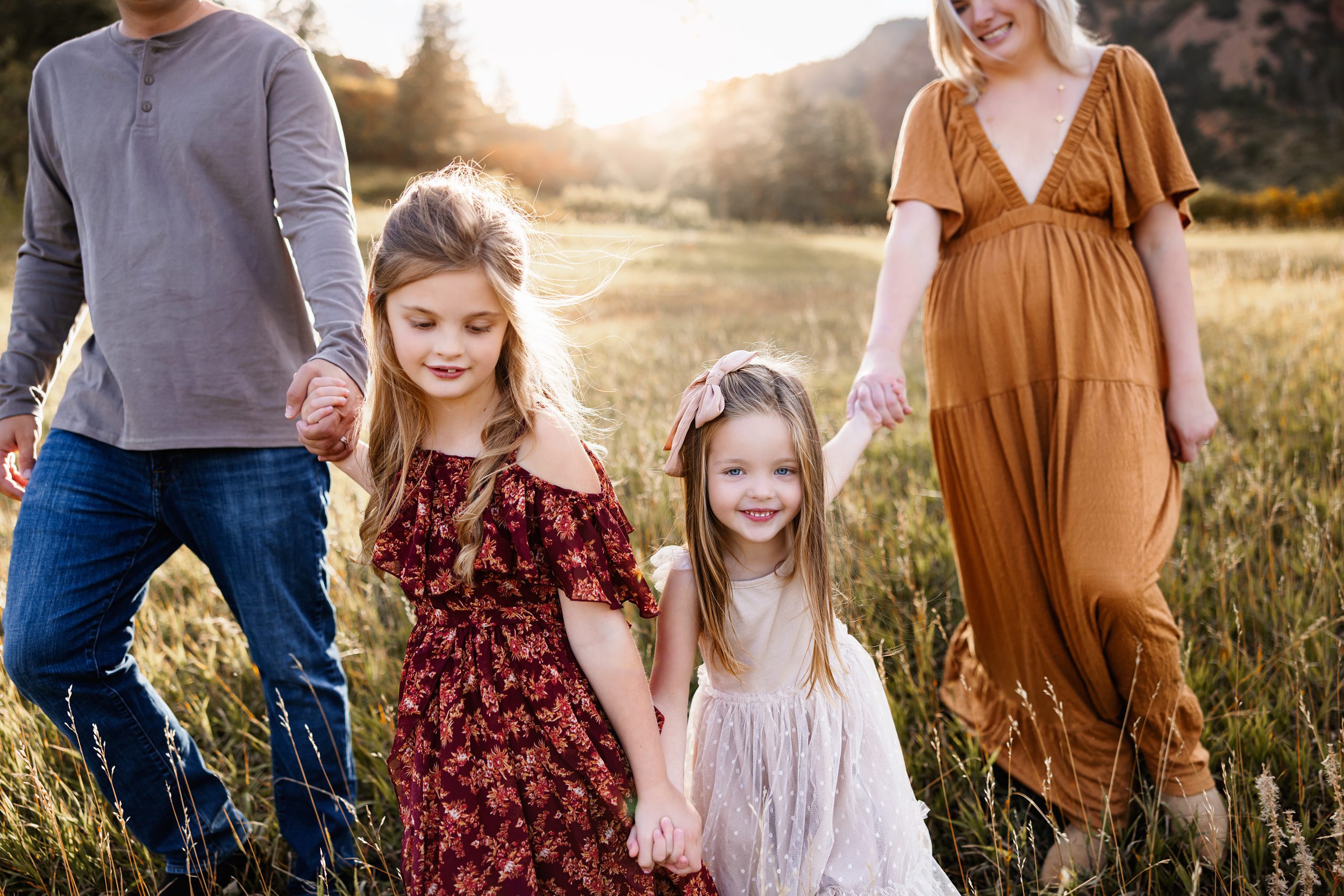 Candid Fall Family Photos Colorado Springs capturing two young sisters holding hands and walking through a sunlit field with their parents during golden hour