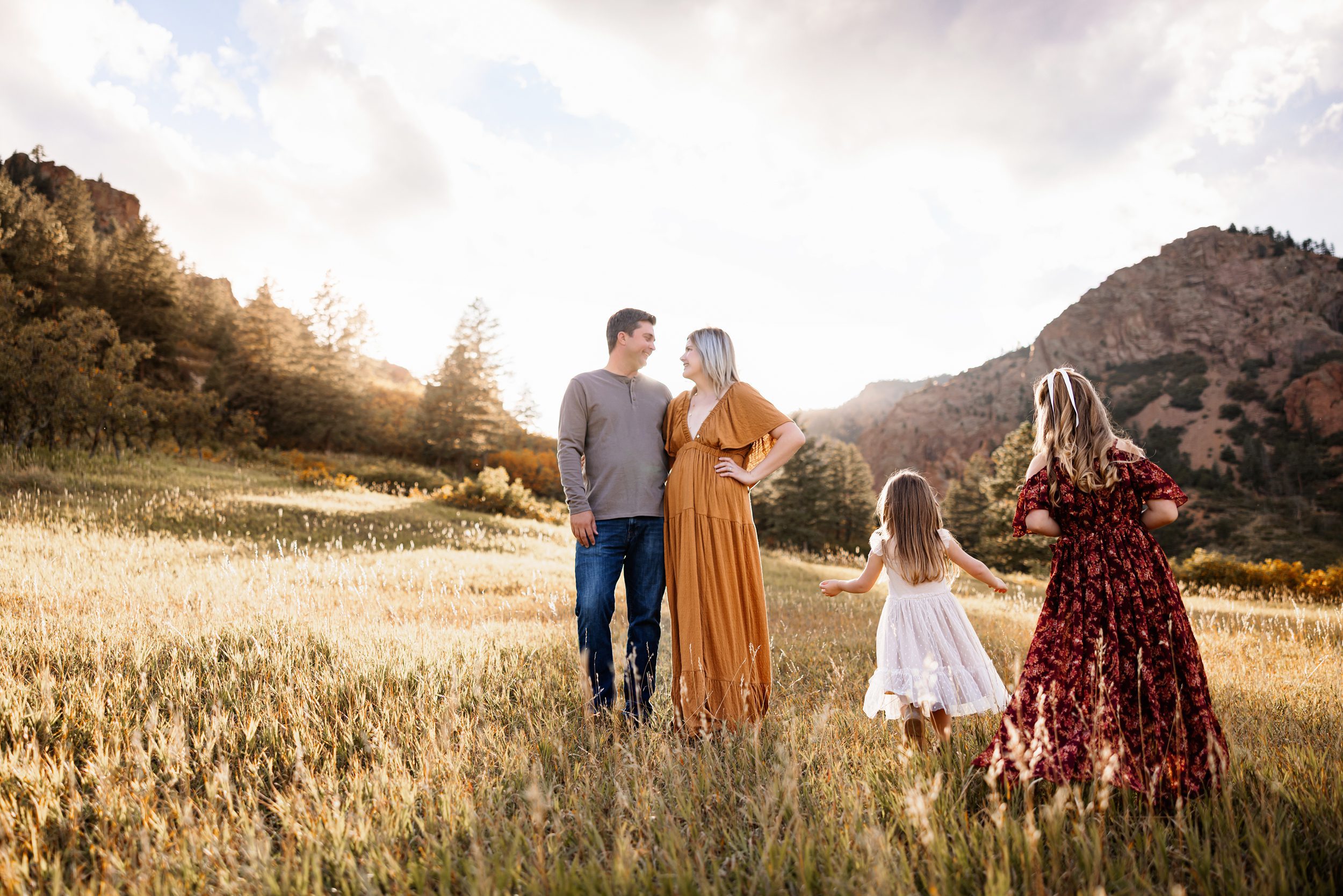 Parents smiling at each other while their daughters run and play through a glowing meadow with mountain views at sunset