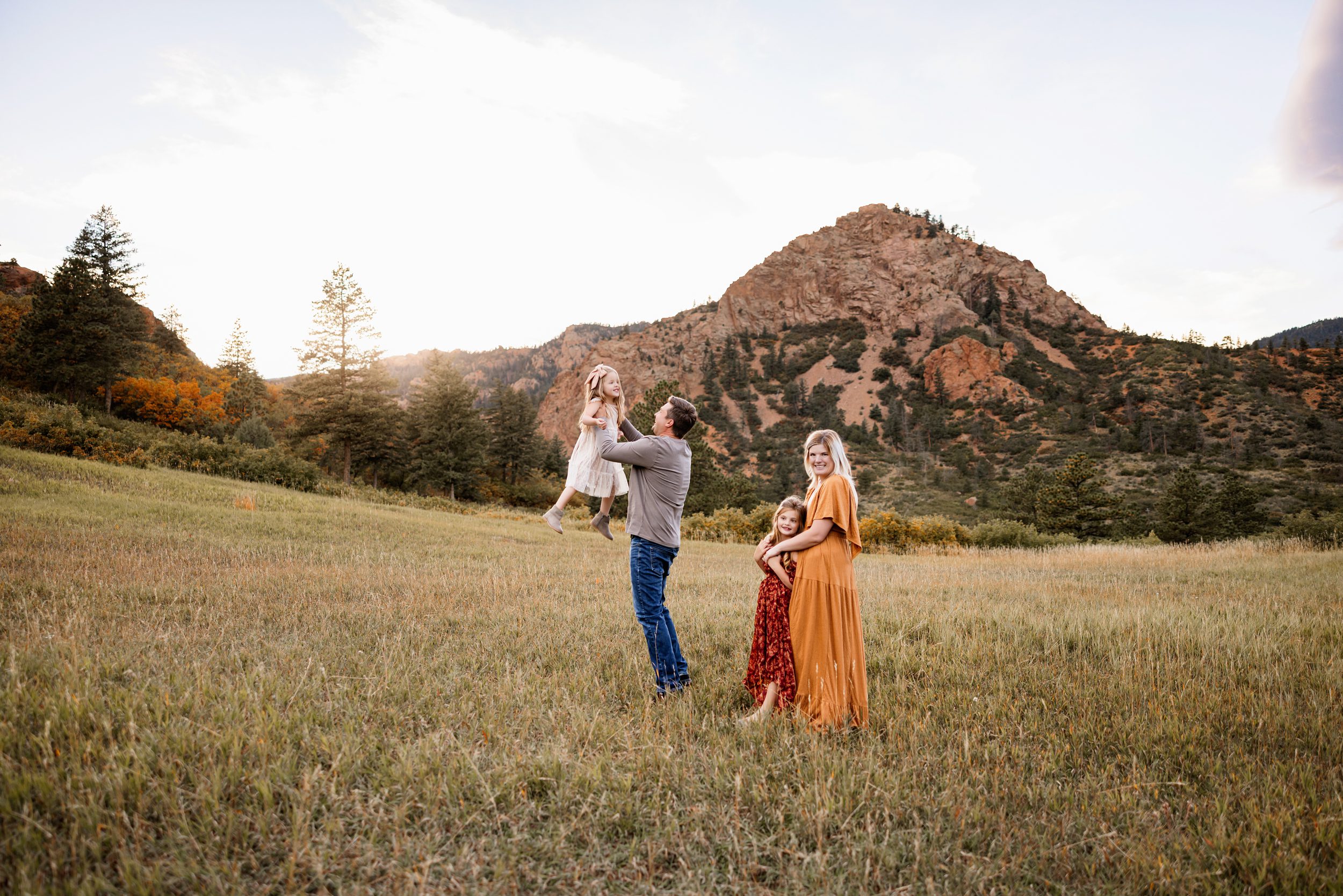 Candid Fall Family Photos Colorado Springs with a dad lifting his daughter in a golden field while mom and sister smile nearby with mountains in the background