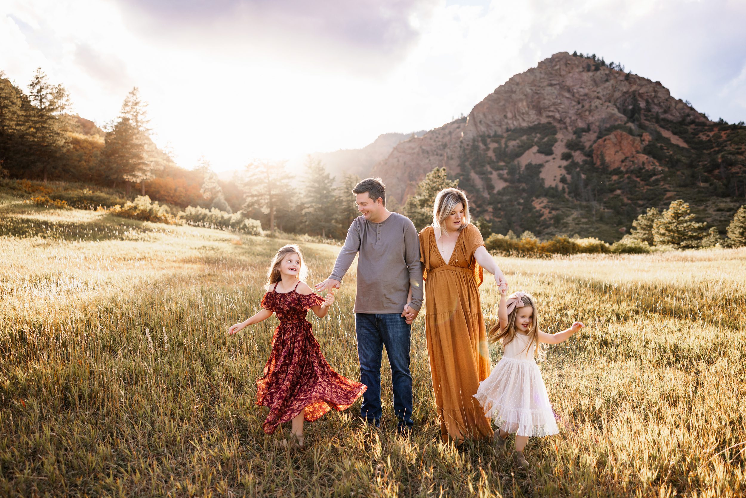 Candid Fall Family Photos Colorado Springs capturing a family holding hands and walking through a glowing field while their daughters twirl and smile in warm sunset light