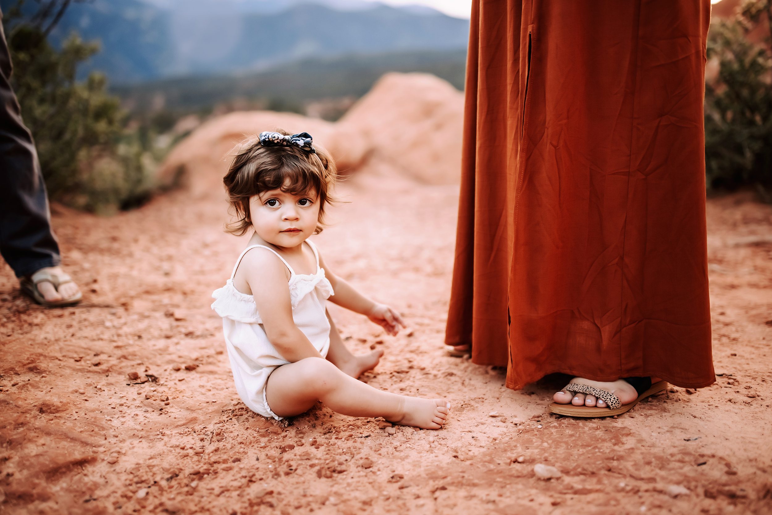 Baby sitting on the red rock dirt at Garden of the Gods wearing a white romper