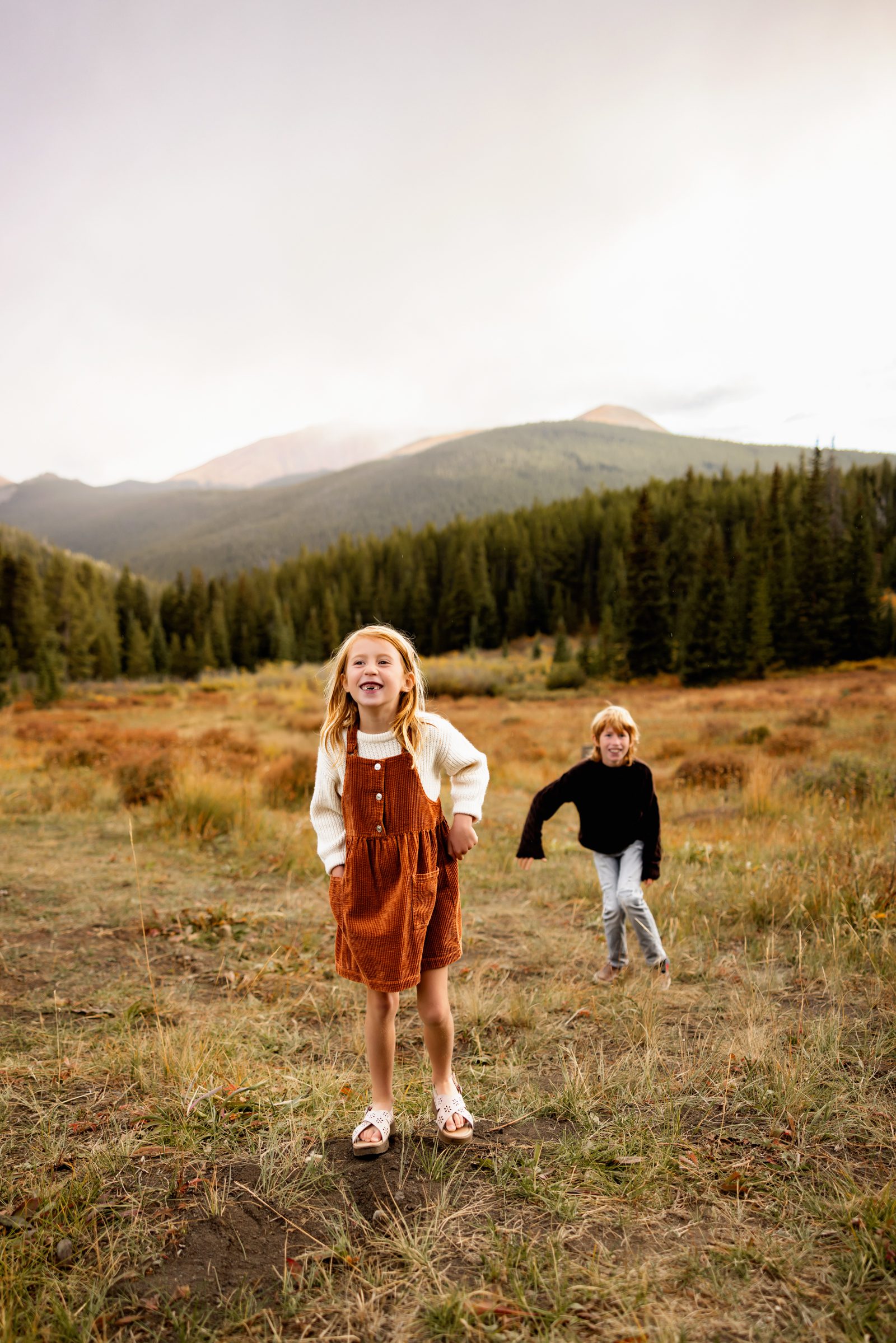 Fall Family Photos in Breckenridge showing playful siblings running through an open meadow with mountains in the background