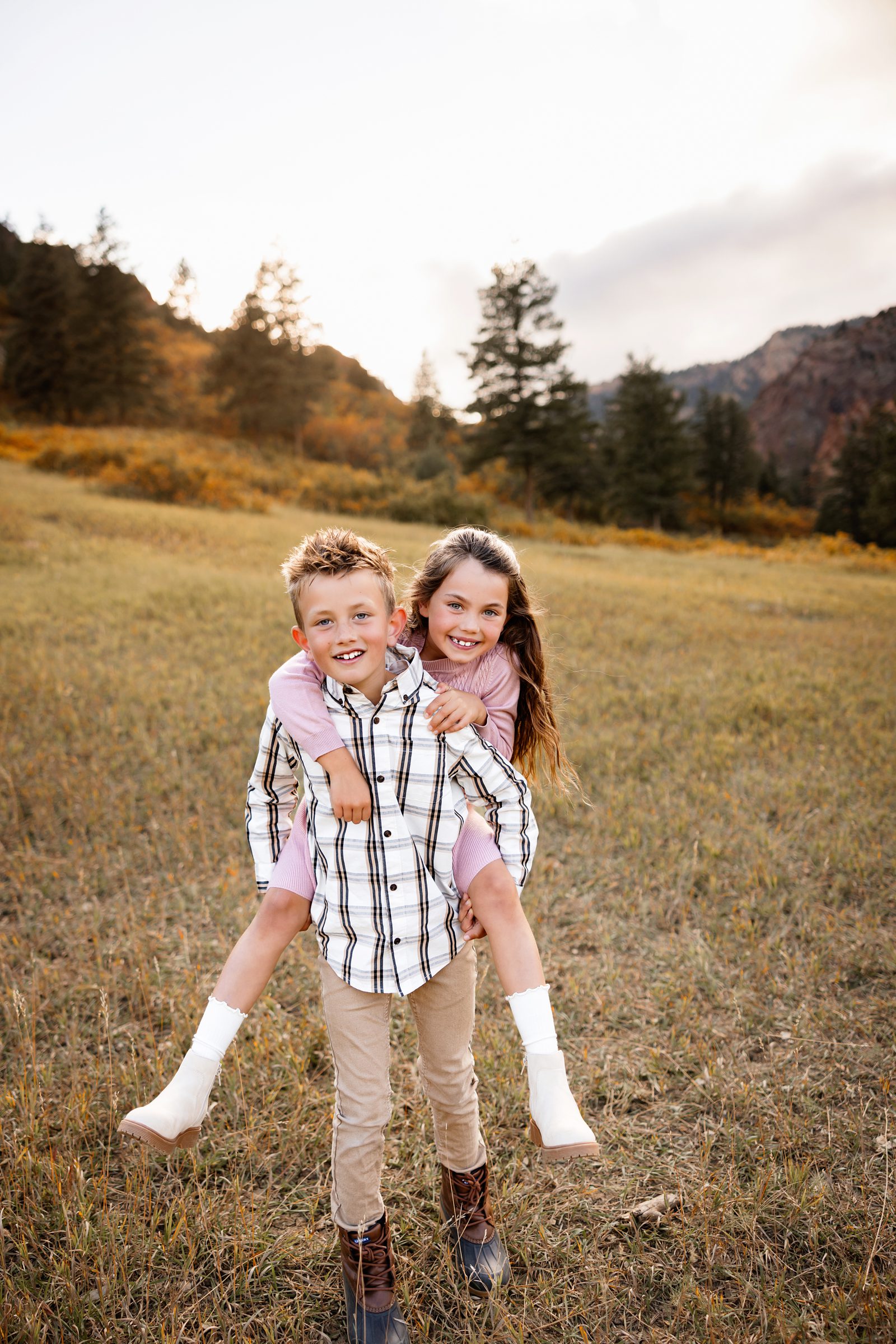 Siblings smiling with piggyback pose in a grassy field with mountain scenery behind them