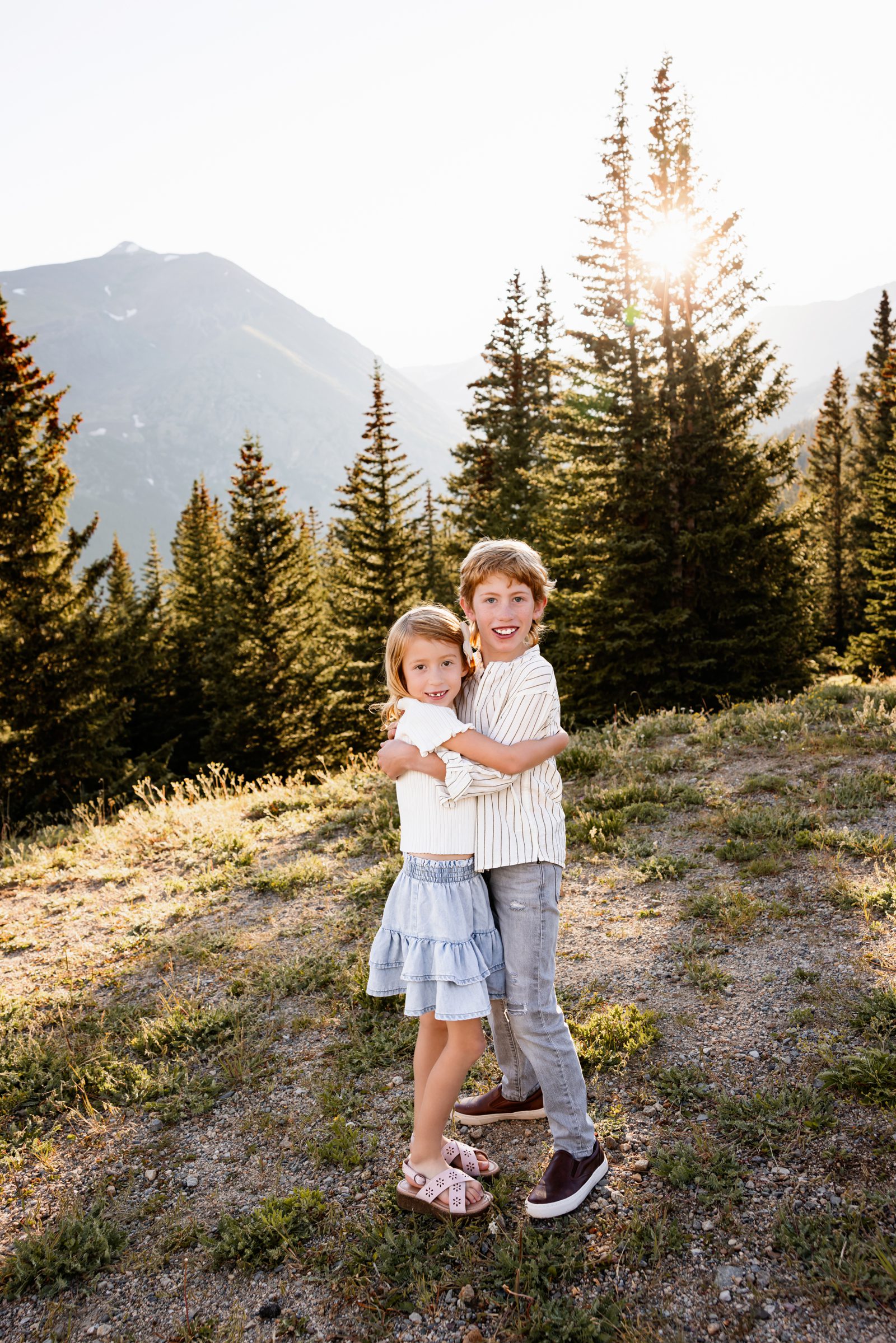 Family Photos in Breckenridge of siblings hugging in a sunlit mountain meadow at Hoosier Pass with pine trees and peaks behind them