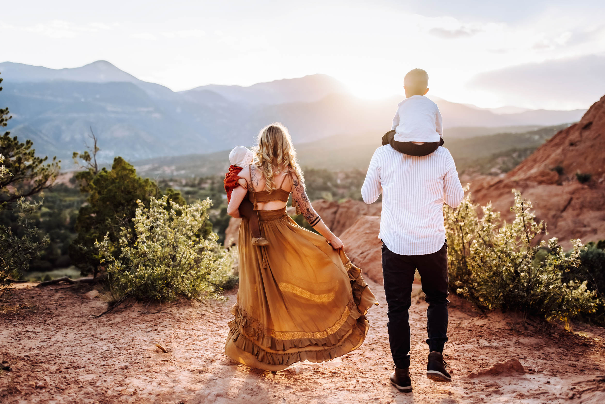 Family walking along a red rock trail at sunset with flowing dress and child on shoulders in Colorado Springs