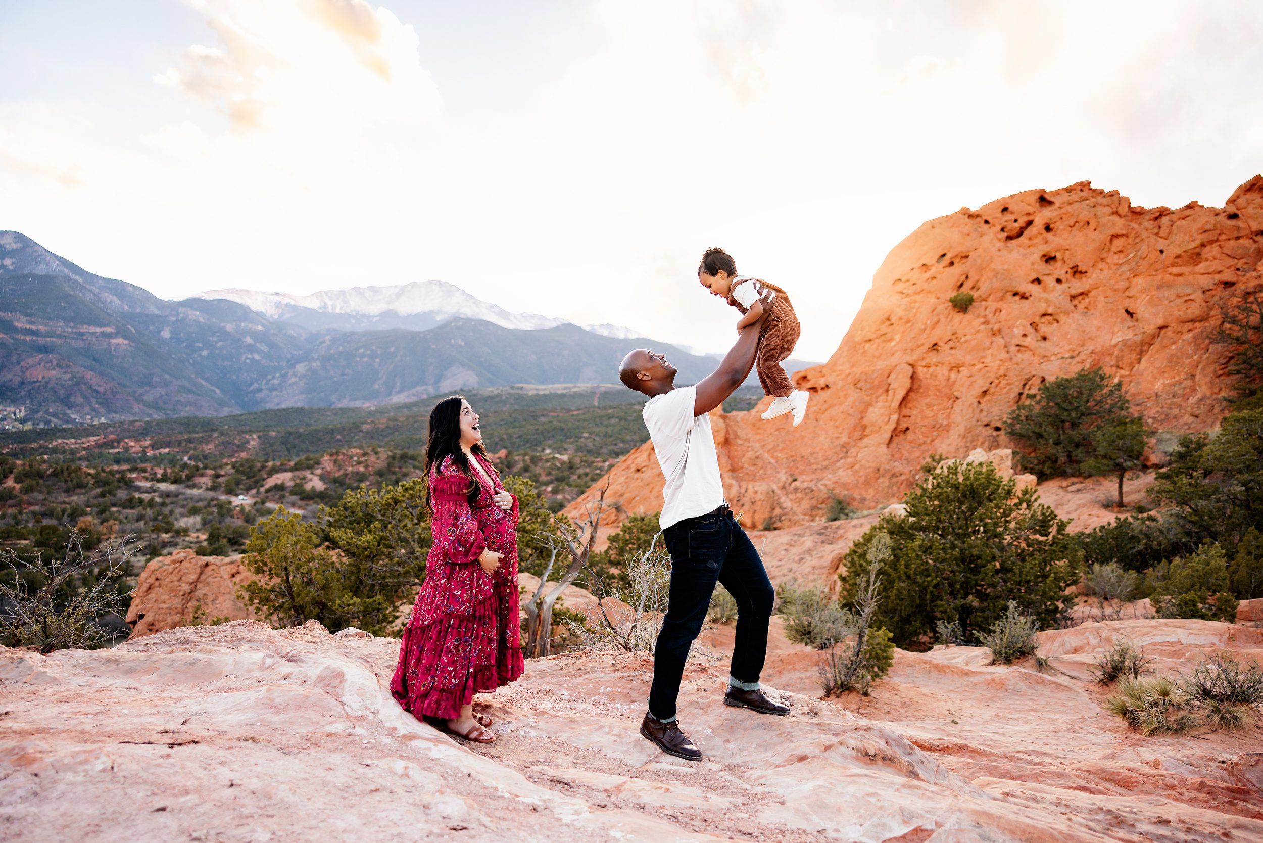 Garden of the Gods family photos of a father lifting his child in the air while mother smiles nearby on red rocks