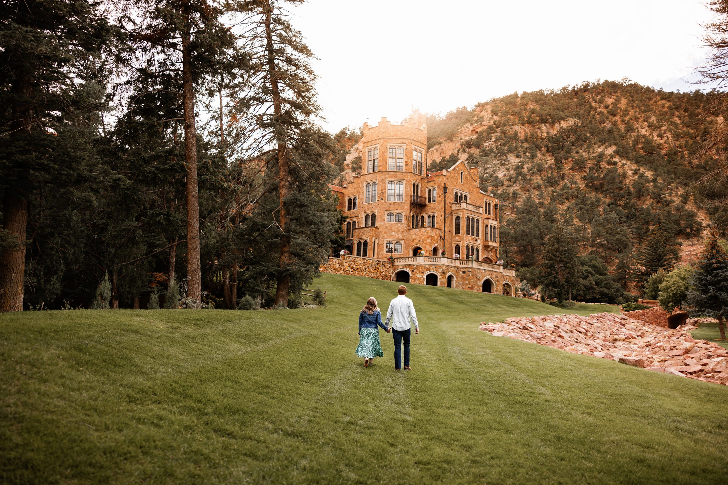 Couple walking hand in hand toward Glen Eyrie castle during golden hour with mountain backdrop in Colorado Spring