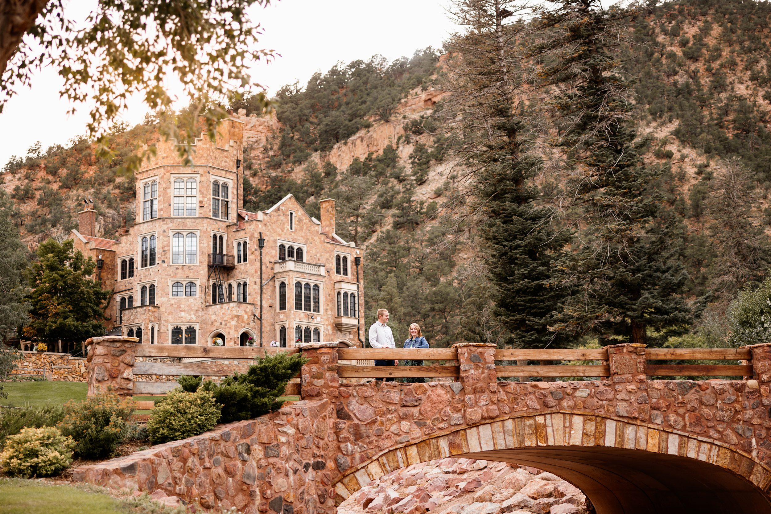 Couple standing on a stone bridge at Glen Eyrie with historic castle and canyon landscape in the background