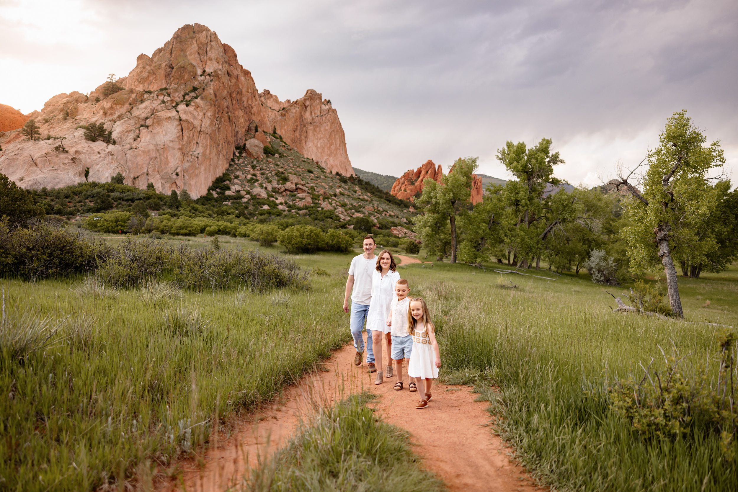 Family walking together through tall grass with dramatic clouds and red rock formations in the background