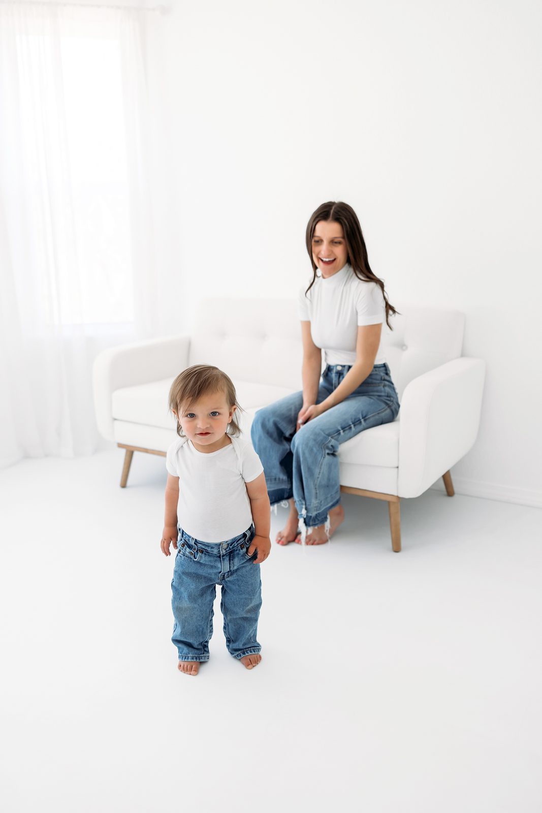 Toddler standing in a white studio while mom sits on a chair in the background