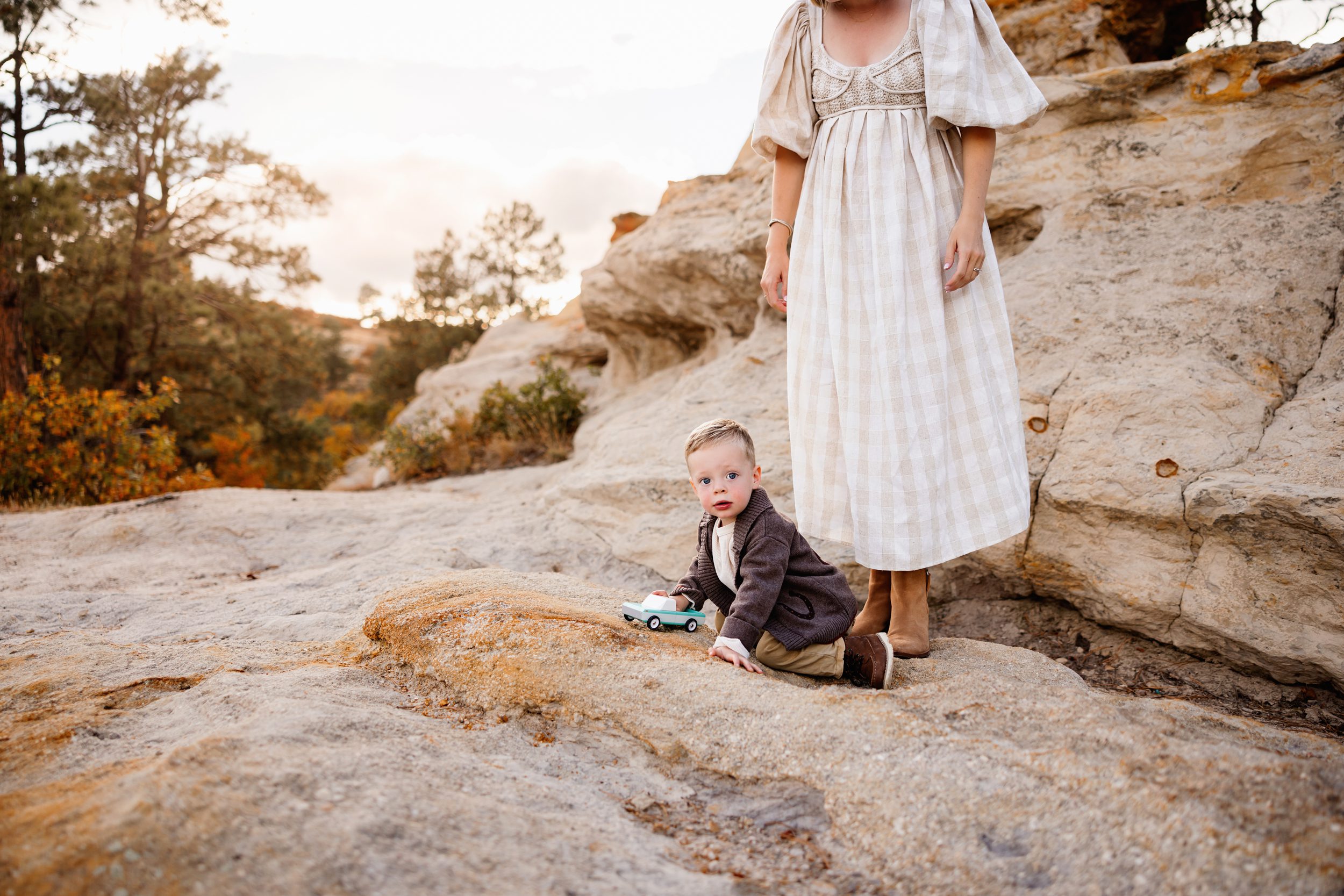 Toddler playing with a toy car on sandstone rocks while mom stands nearby in a flowing dress during a relaxed fall family session