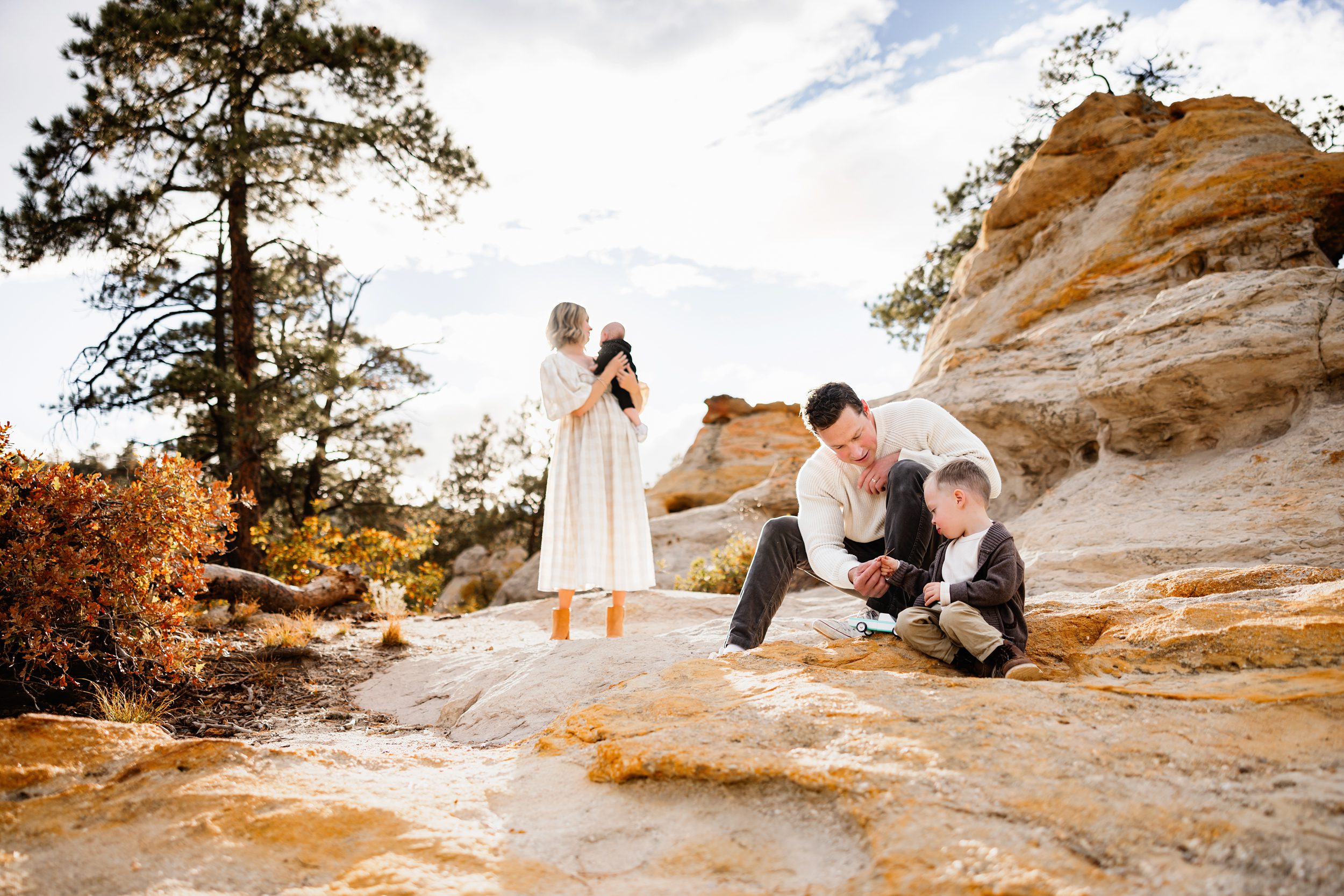 When to schedule your fall photos in Colorado with a candid moment of dad and toddler playing on rocky terrain while mom holds baby in the background