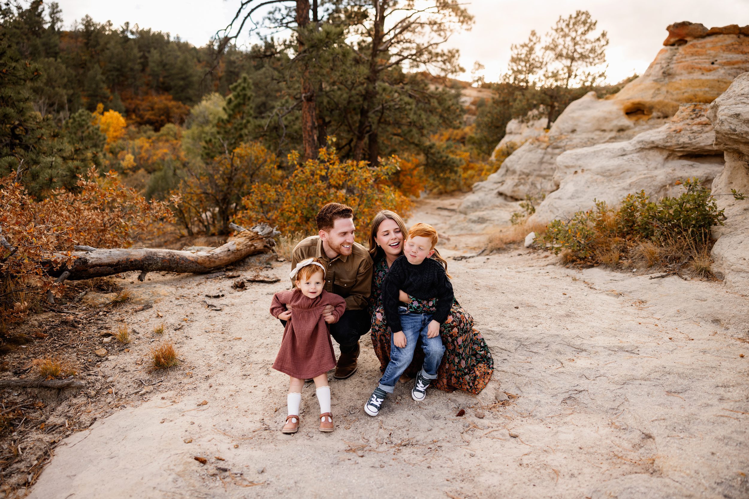 Photo session with toddler in Colorado Springs capturing a playful family moment with parents tickling their kids on a sandy trail surrounded by fall colors