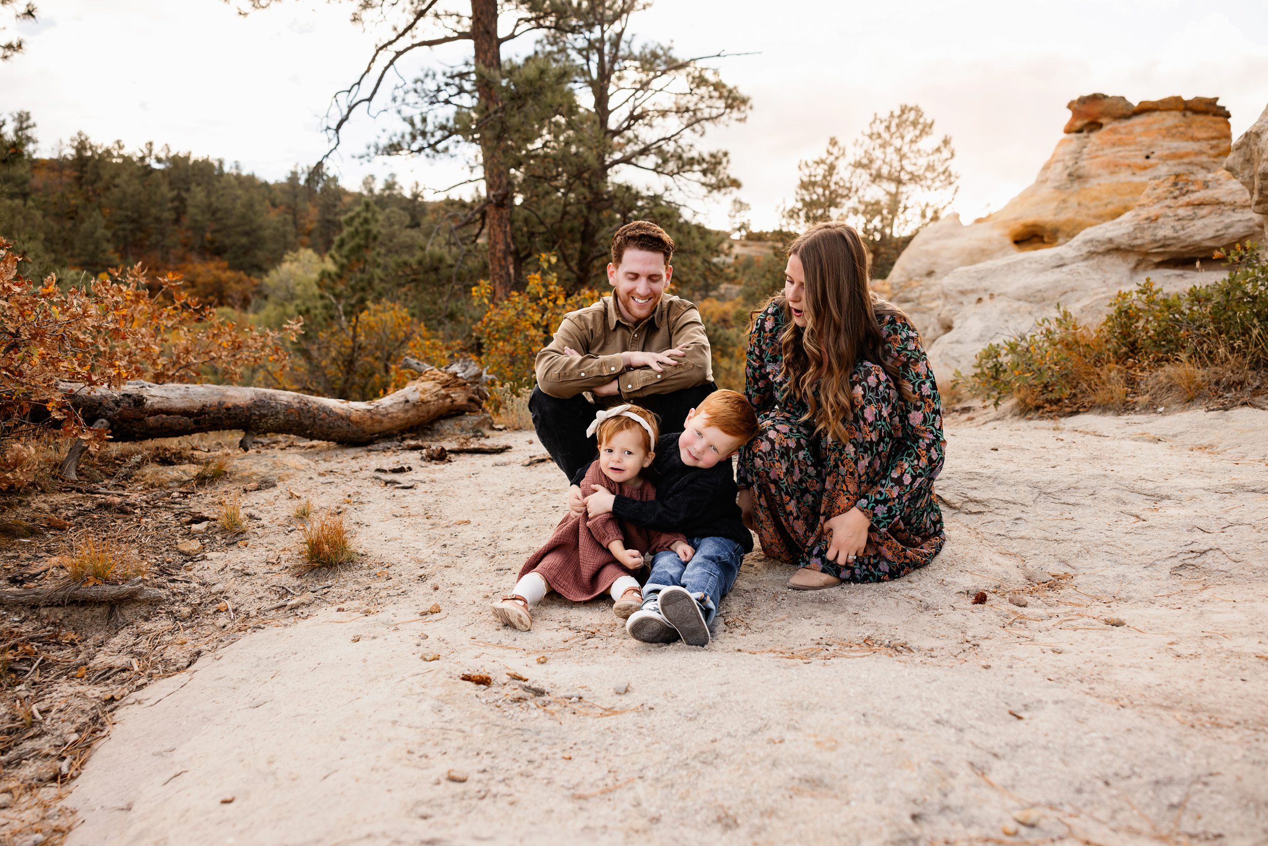 Close candid of siblings sitting together while parents smile nearby in a relaxed outdoor setting