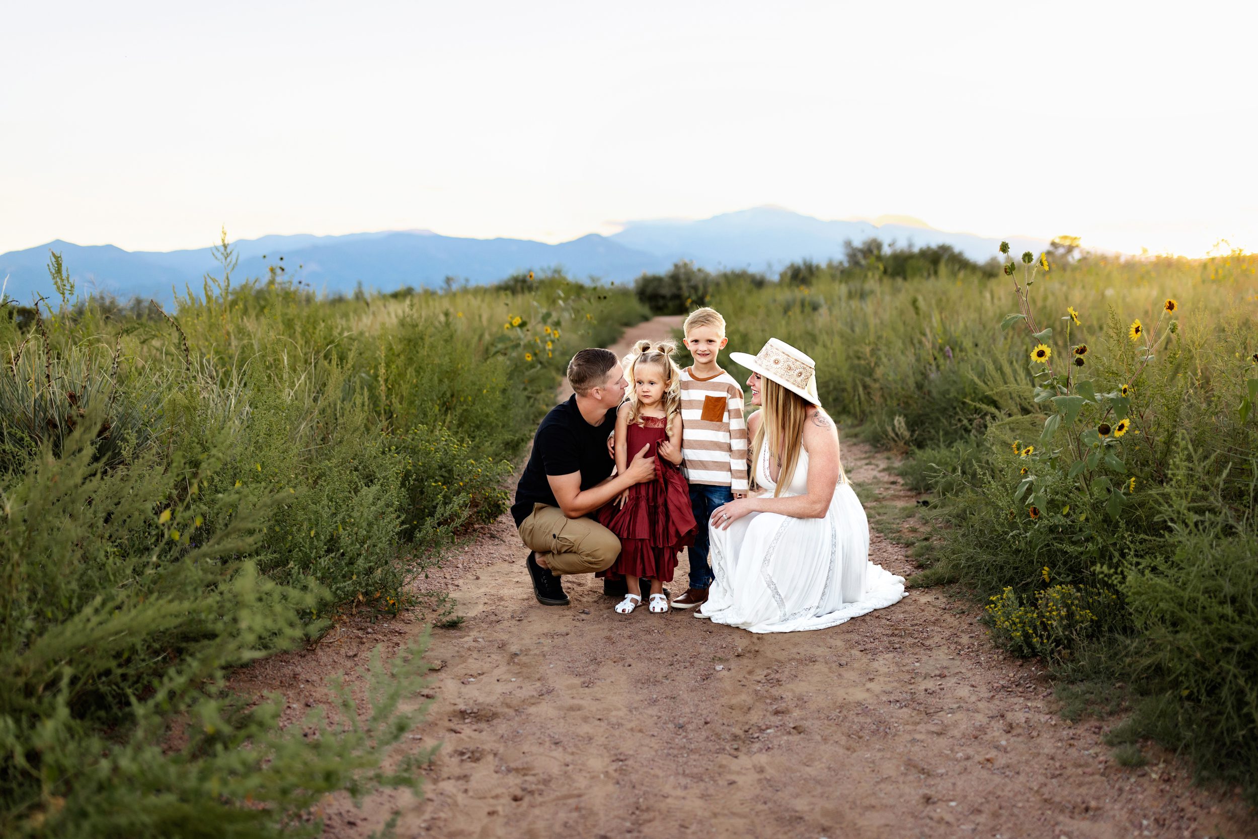 Family of four smiling together on a dirt path surrounded by wildflowers and open space