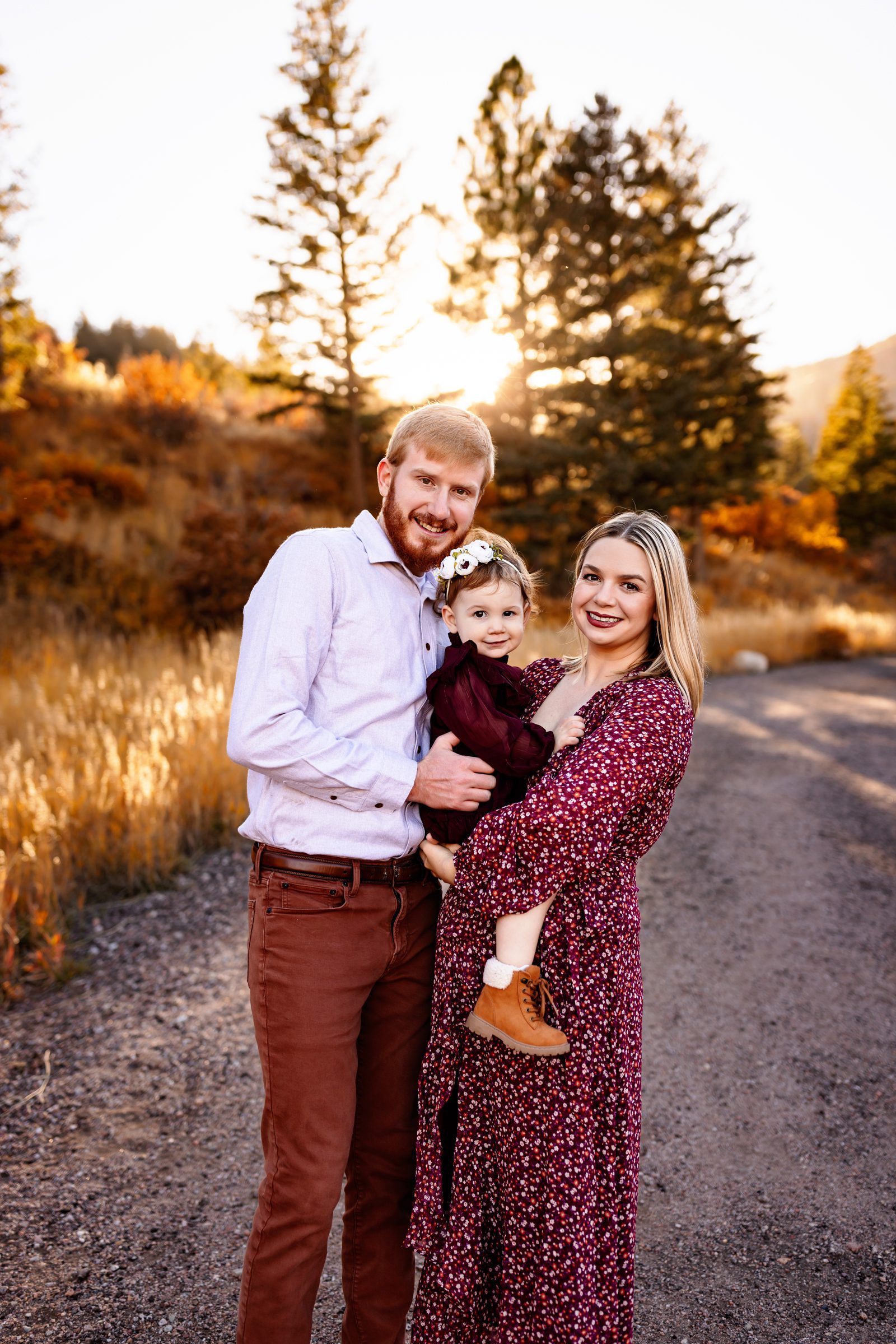 Smiling parents holding their toddler in golden light with colorful trees in the background