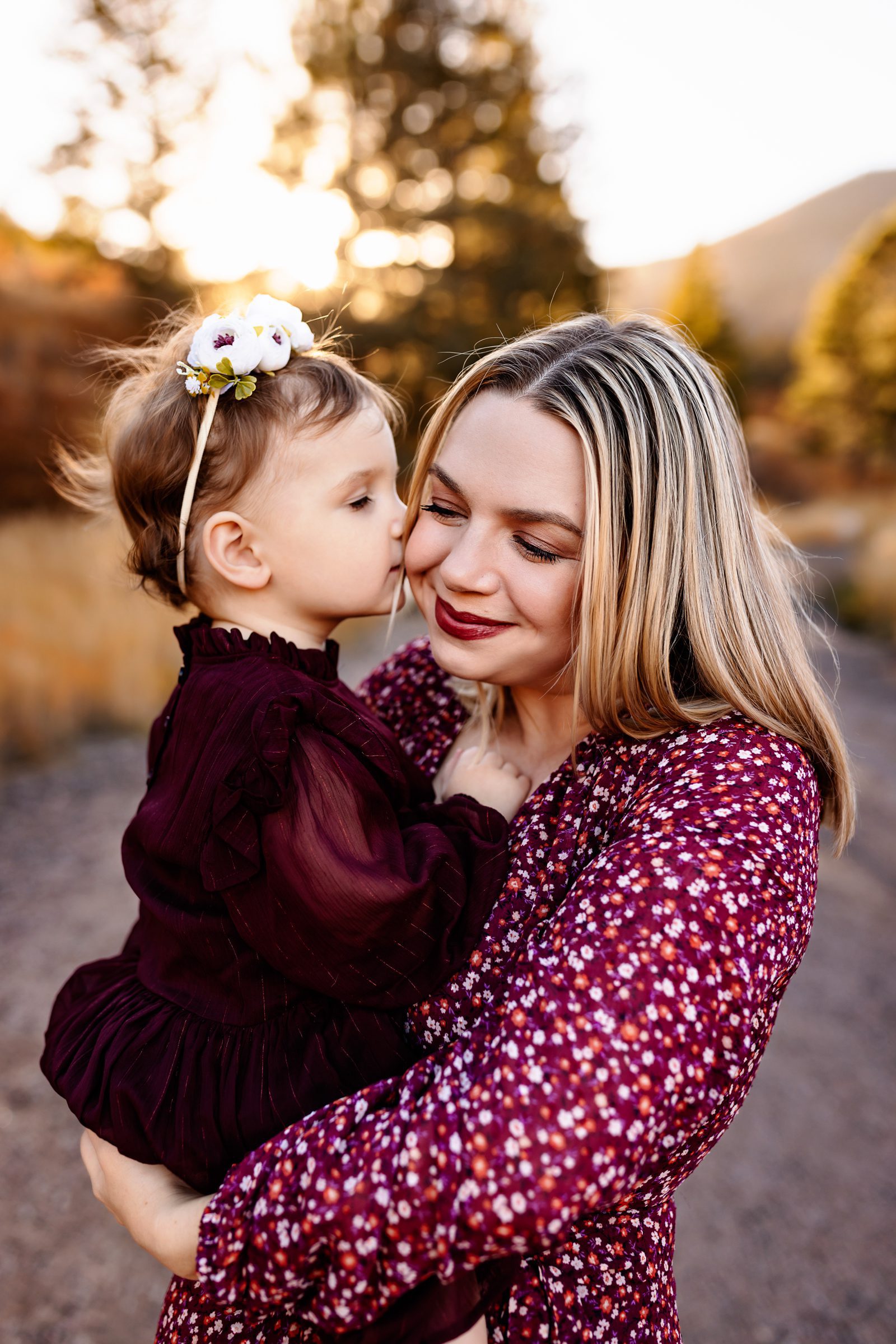 Close up of a mother holding her toddler as the child kisses her cheek in warm sunset light