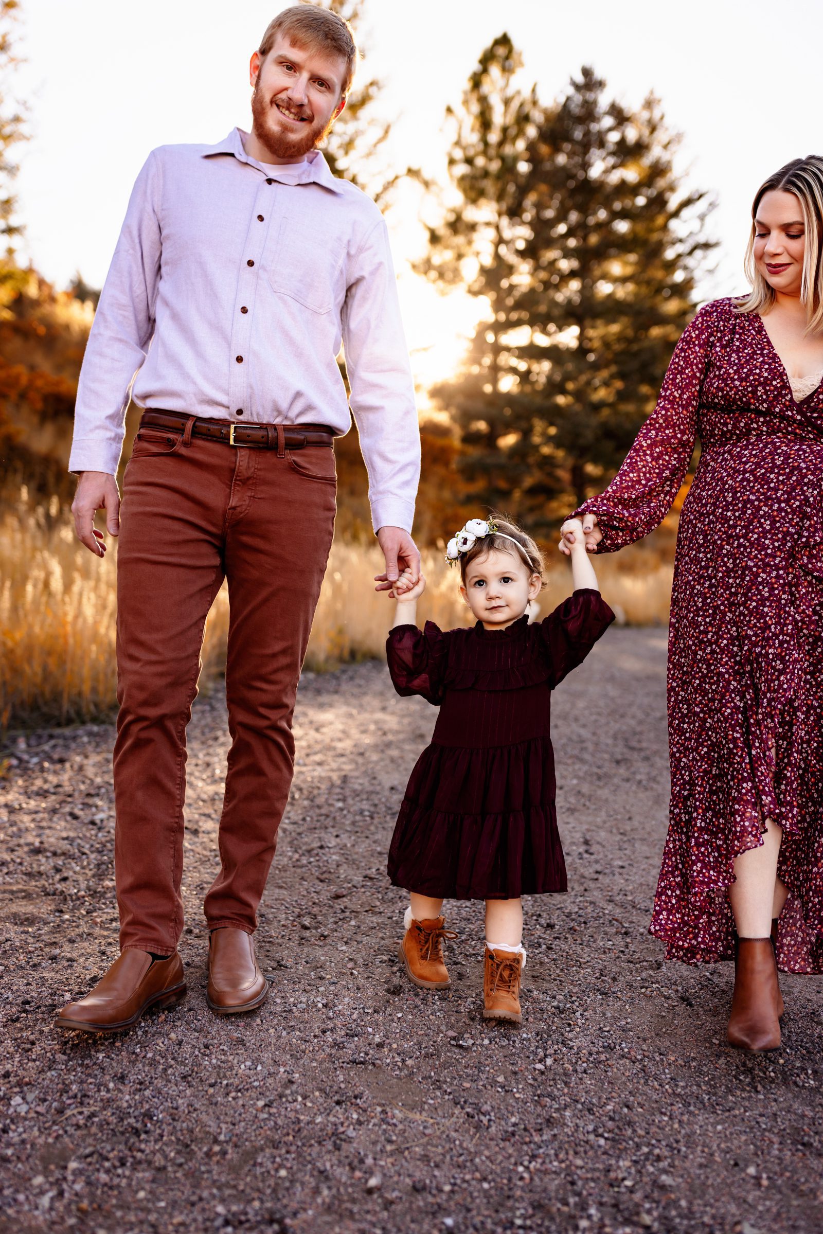 Parents holding their toddler’s hands while walking along a scenic path with warm autumn tones