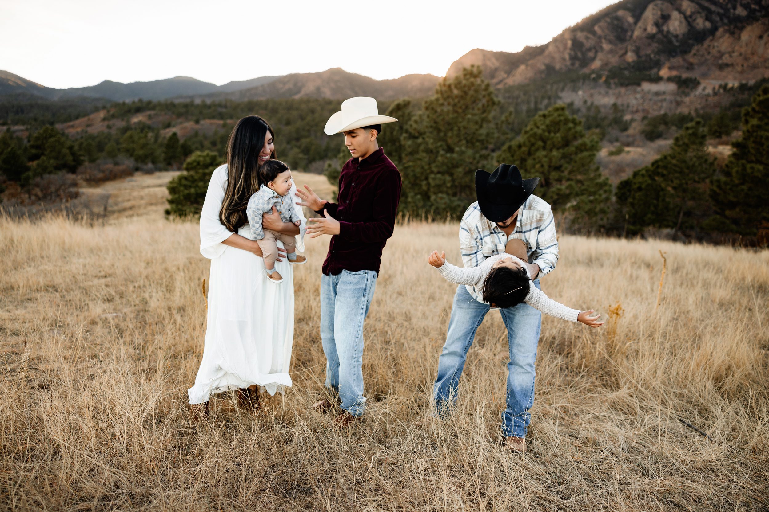 Candid family moment in a golden field with parents and children laughing and playing, set against mountain views at sunset