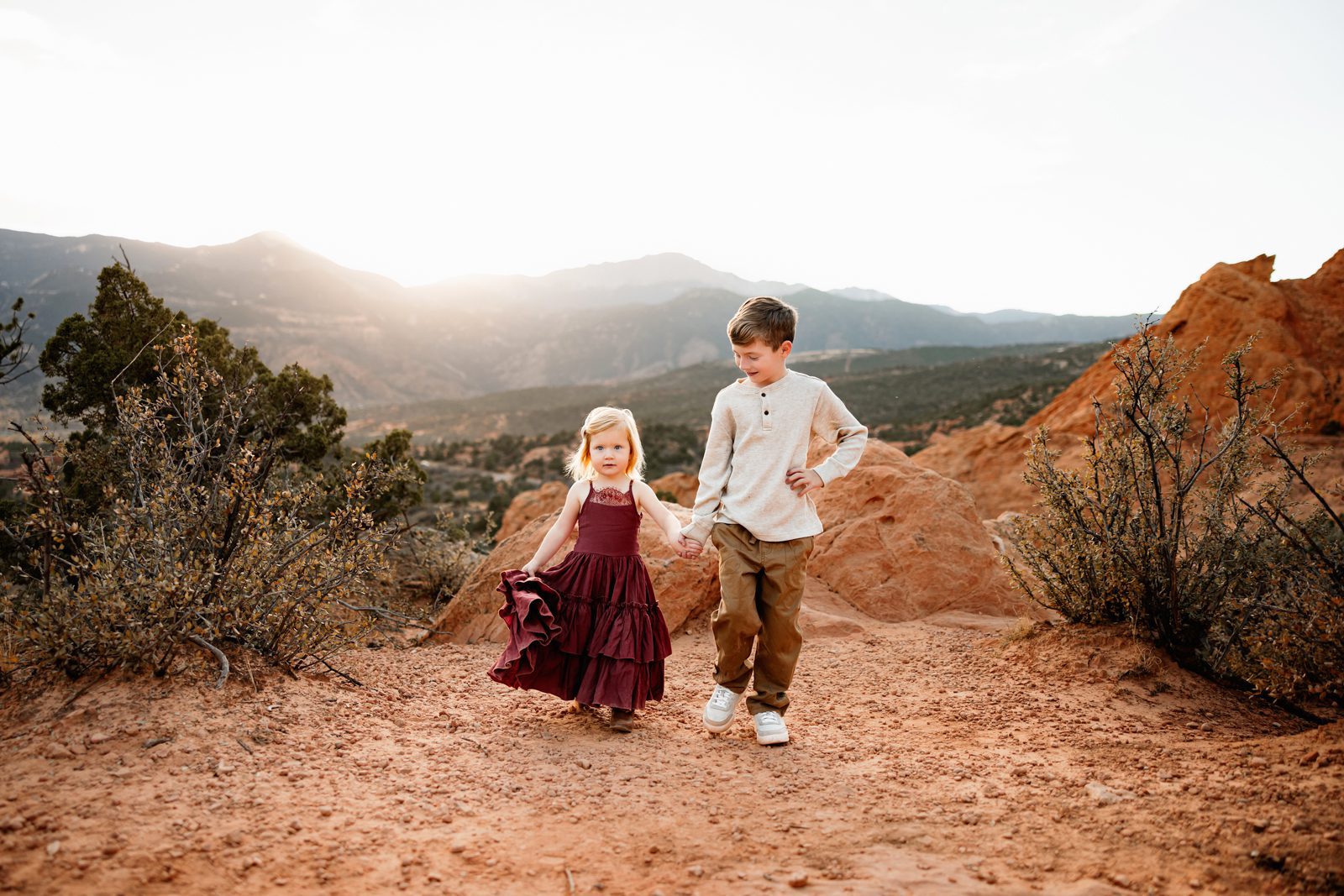Siblings walking together on a dirt path surrounded by red rocks and scenic mountain views during golden hour