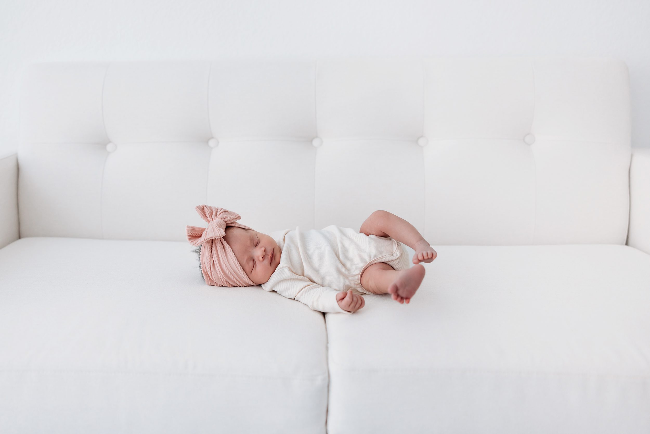 Newborn session in Colorado Springs with baby girl sleeping on a white couch wearing a soft pink bow headband