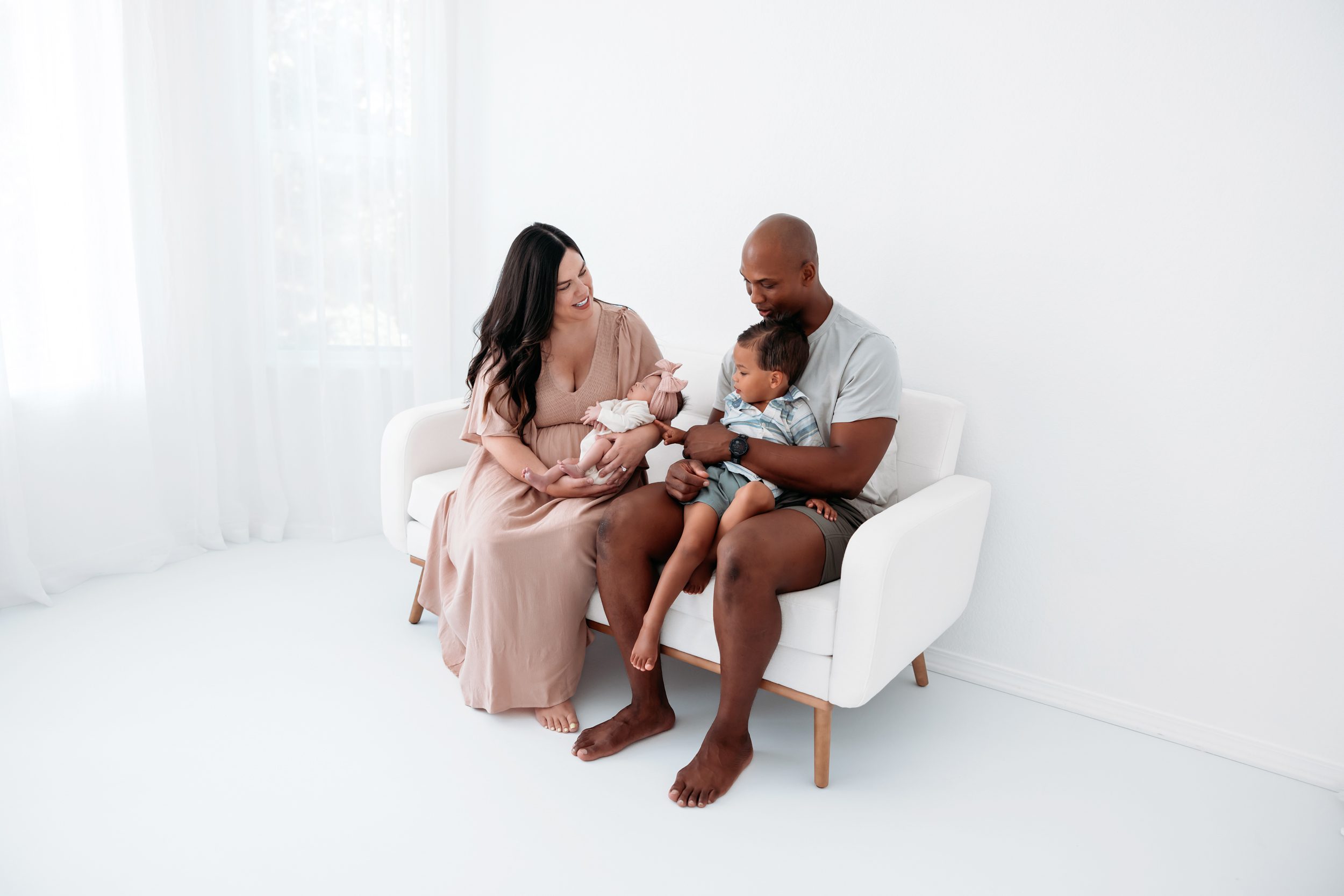 Parents sitting together in a bright white studio holding their newborn and toddler for a relaxed family portrait