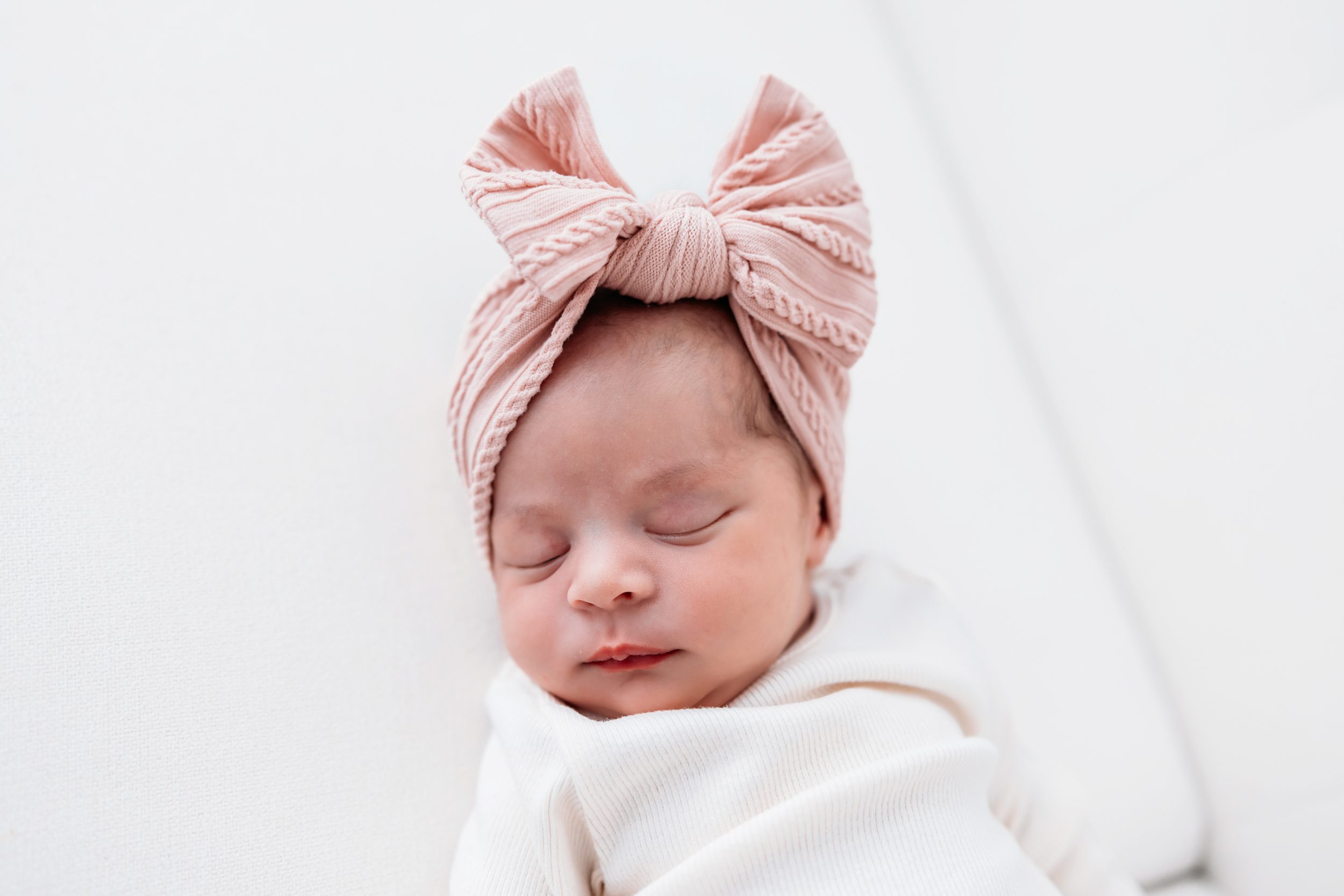Close-up of a sleeping newborn wrapped in a cream swaddle with a pink bow headband in a minimal studio