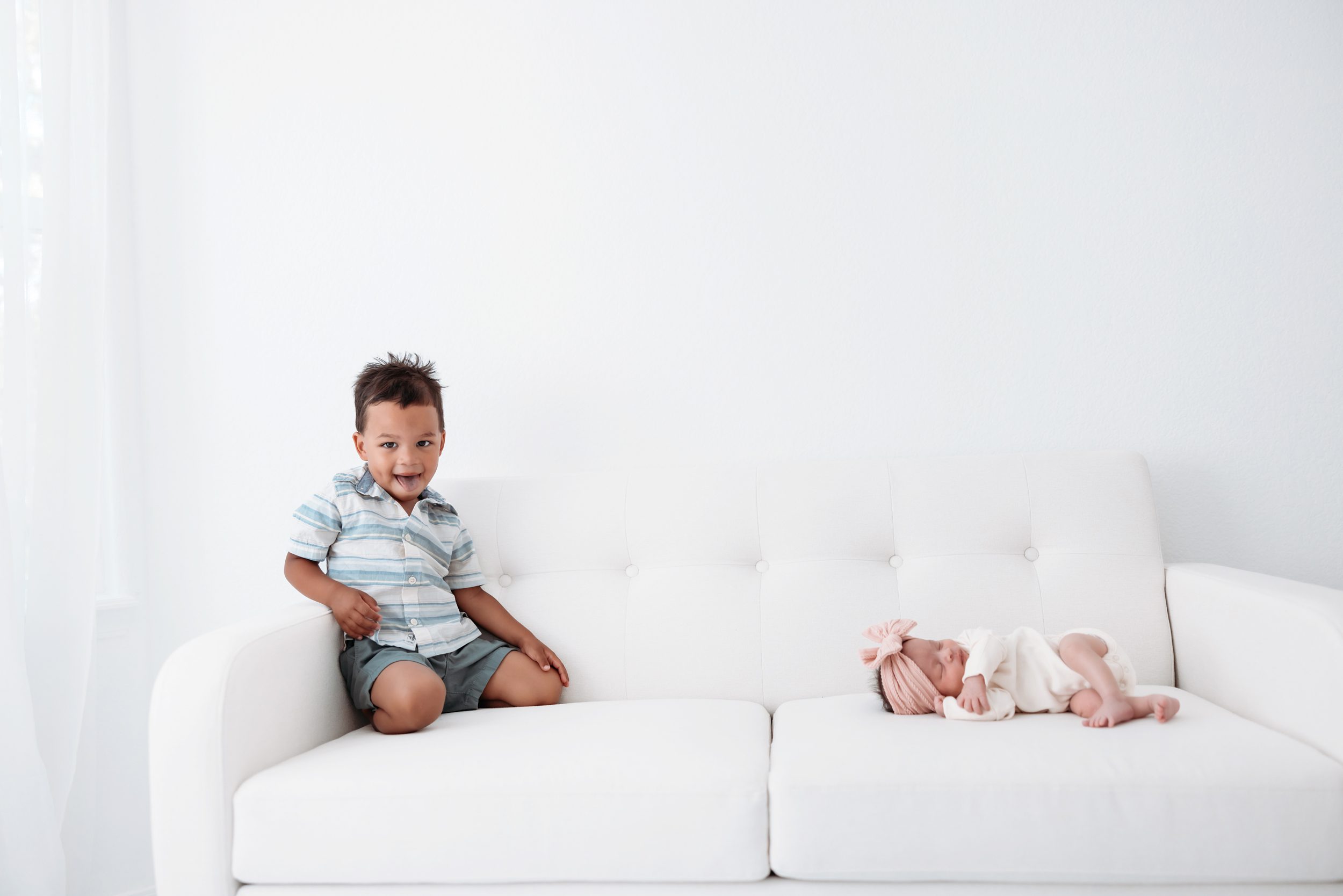 Playful toddler leaning over a white couch smiling at sleeping newborn sibling during a studio session