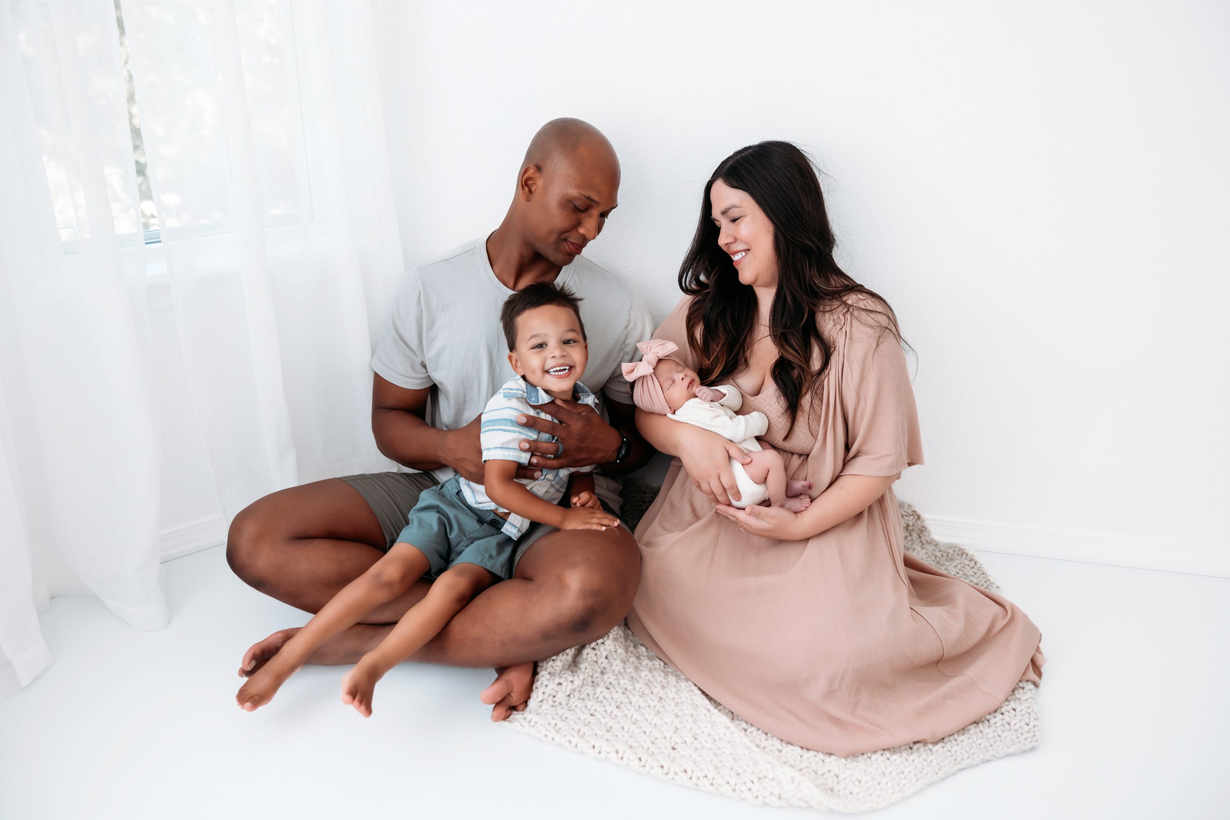 Candid family moment with parents sitting on the floor holding newborn while toddler smiles beside them in a bright studio
