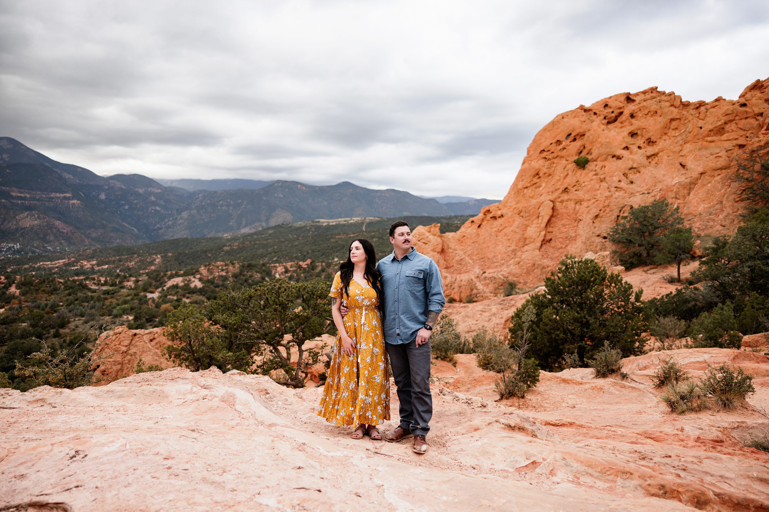 Couple standing together on sandstone rocks with dramatic Colorado landscape and overcast sky creating soft, moody light